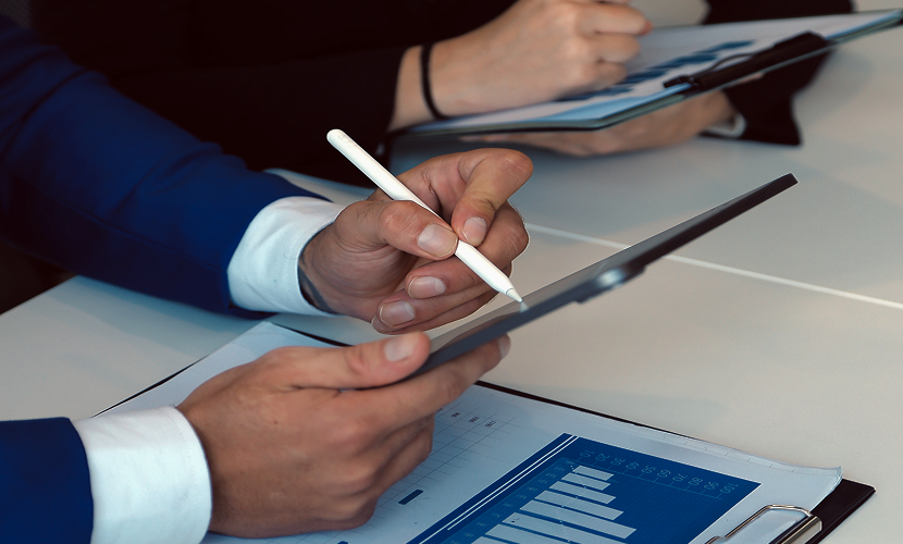 Two people sitting at a table working with tablets and documents, one is writing on a tablet with a stylus, and charts and graphs are spread across the table.