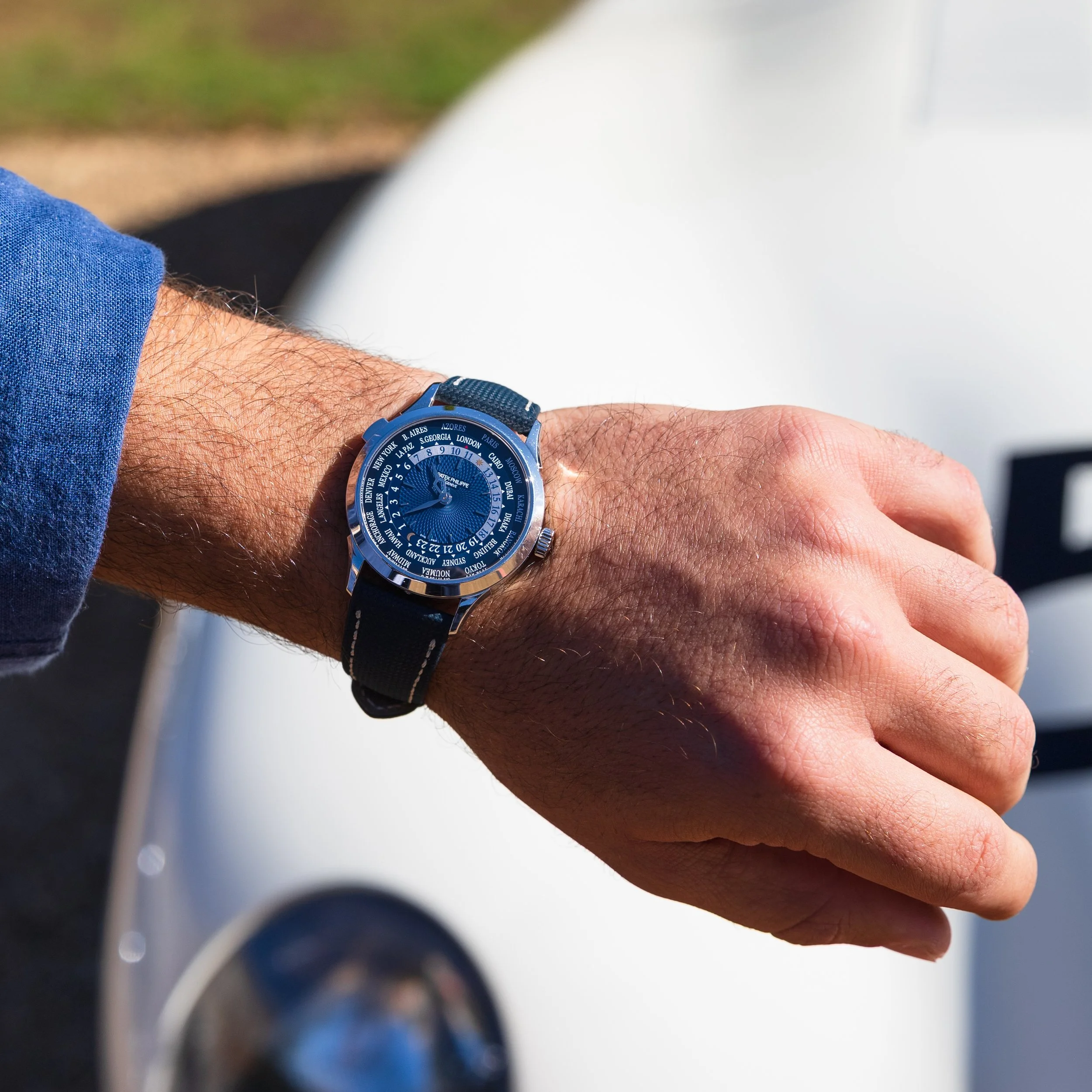 A person's left wrist wearing a blue world time watch with a dark strap, in front of a white vehicle and an outdoor setting.