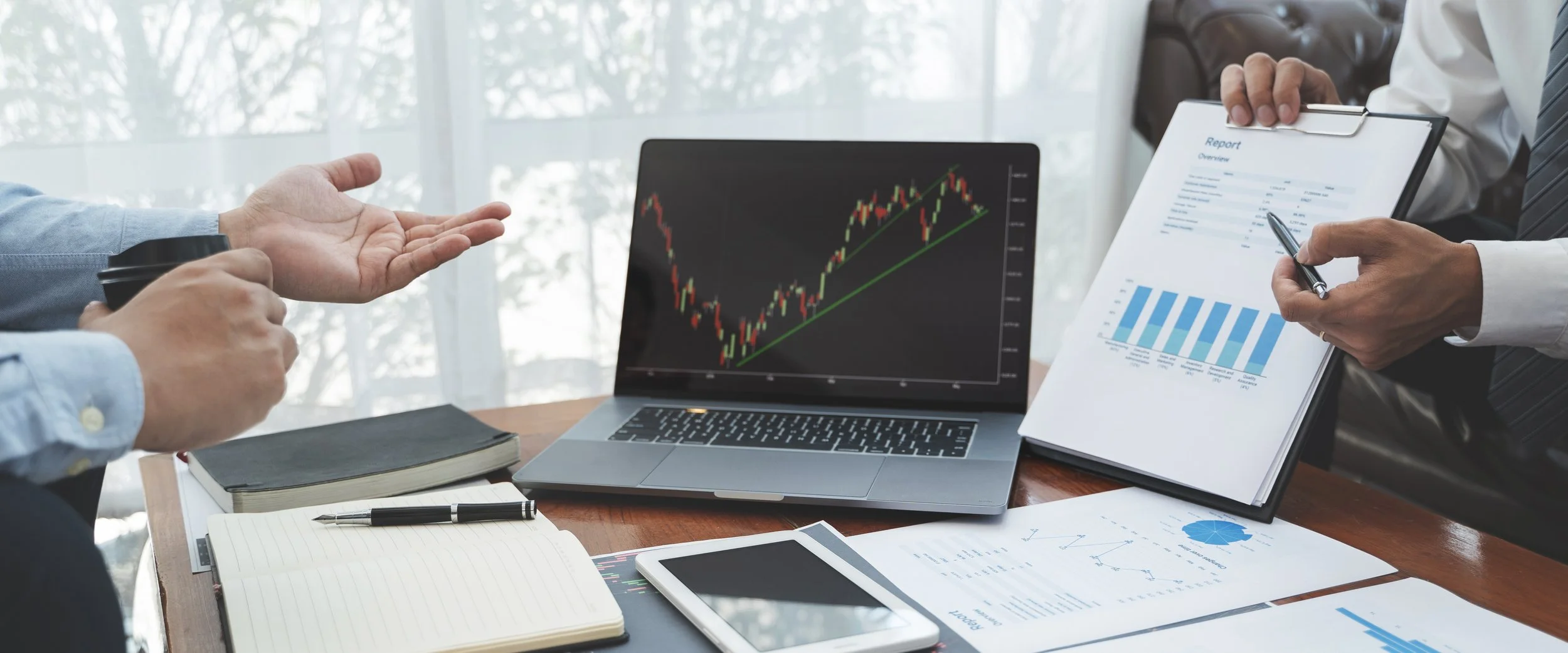 Two professionals discussing financial data at a meeting. One is holding a to-go coffee, and the other is holding a report with charts and graphs. A laptop displaying a stock market chart is on the table, along with notebooks, a pen, and a tablet.