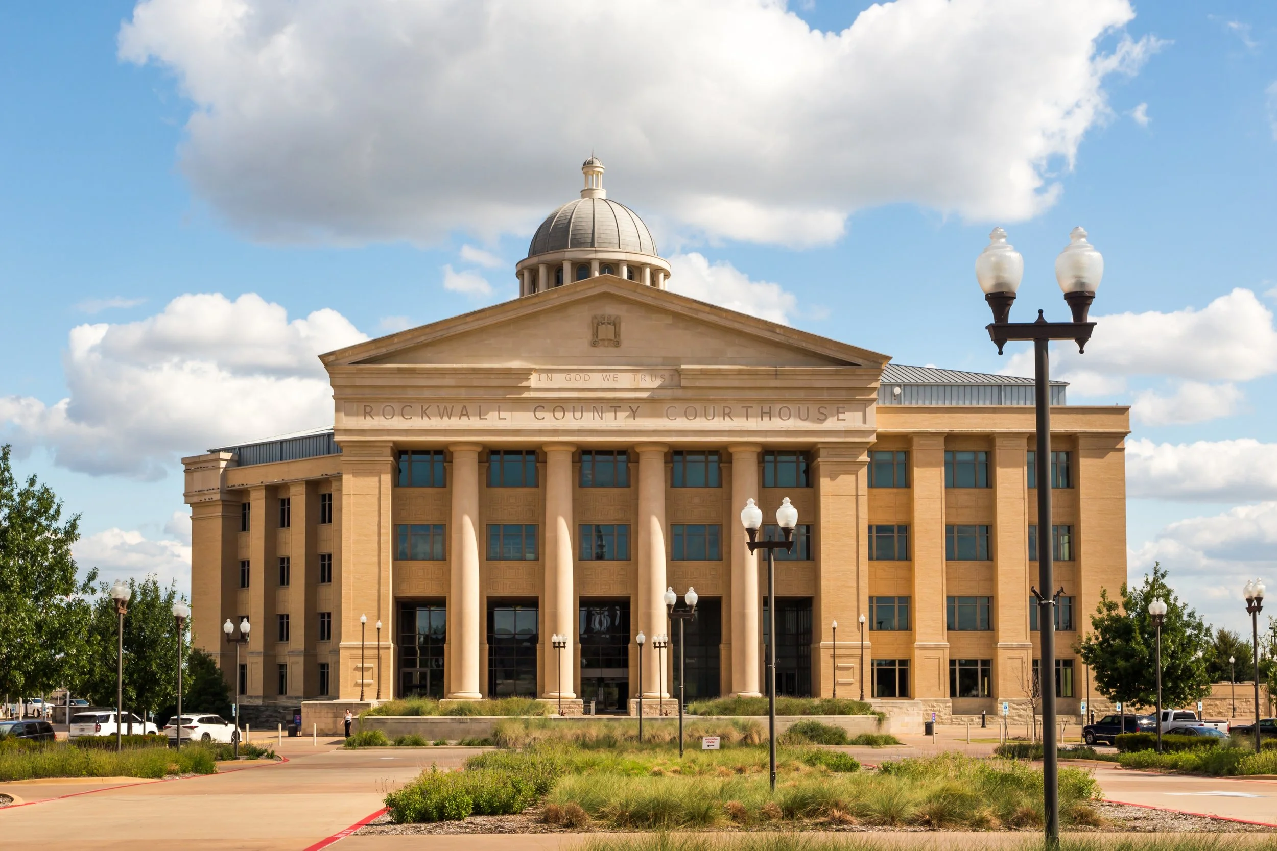 The Rockwall County Courthouse, a historic building with classical architecture, stone facade, tall columns, and a dome, is situated in a landscaped area under a partly cloudy sky.