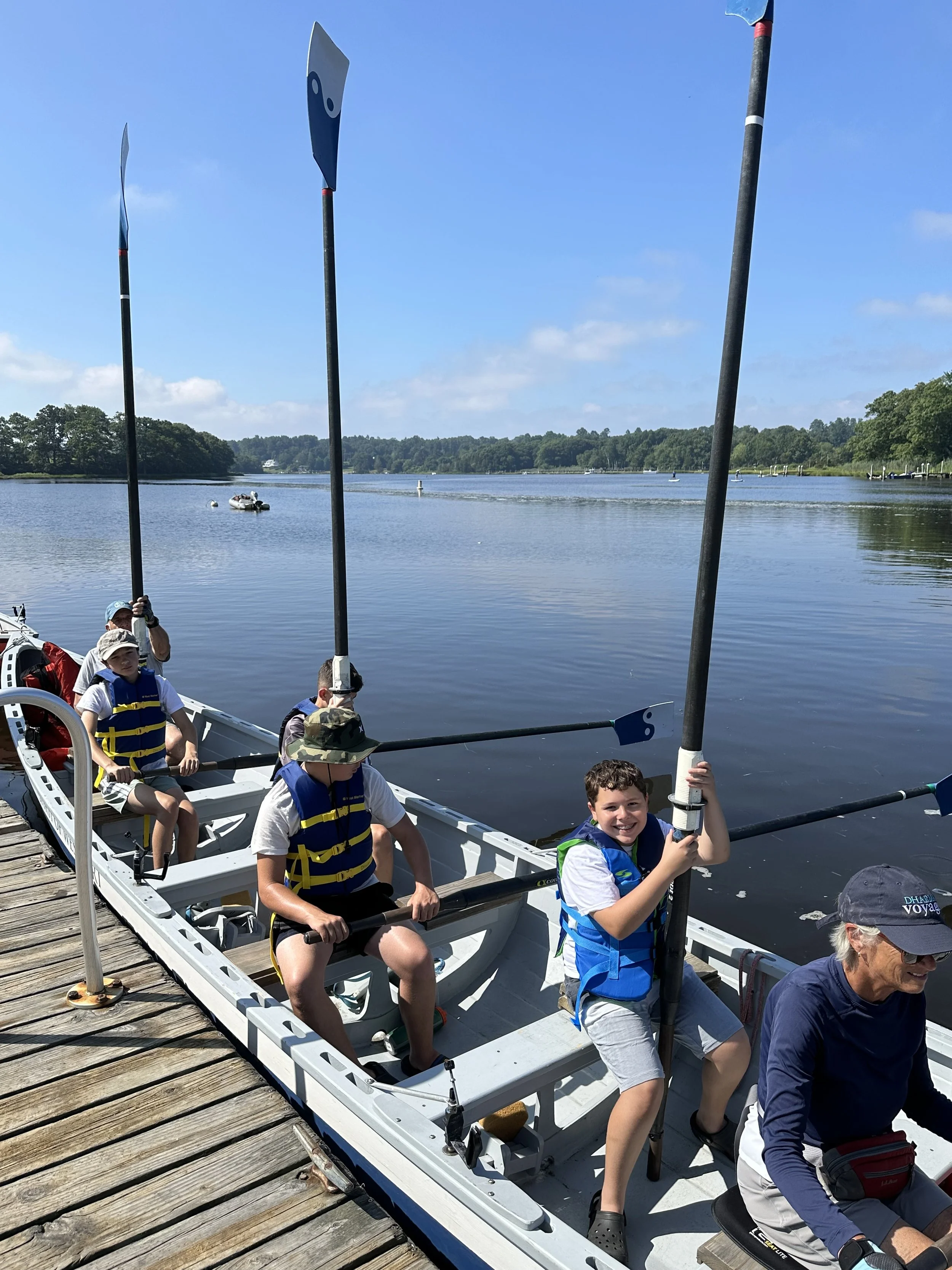 Group of children and an adult in a boat on a lake, holding black paddles with blue blades, near a wooden dock, with a background of calm water and tree-lined shoreline under a partly cloudy sky.