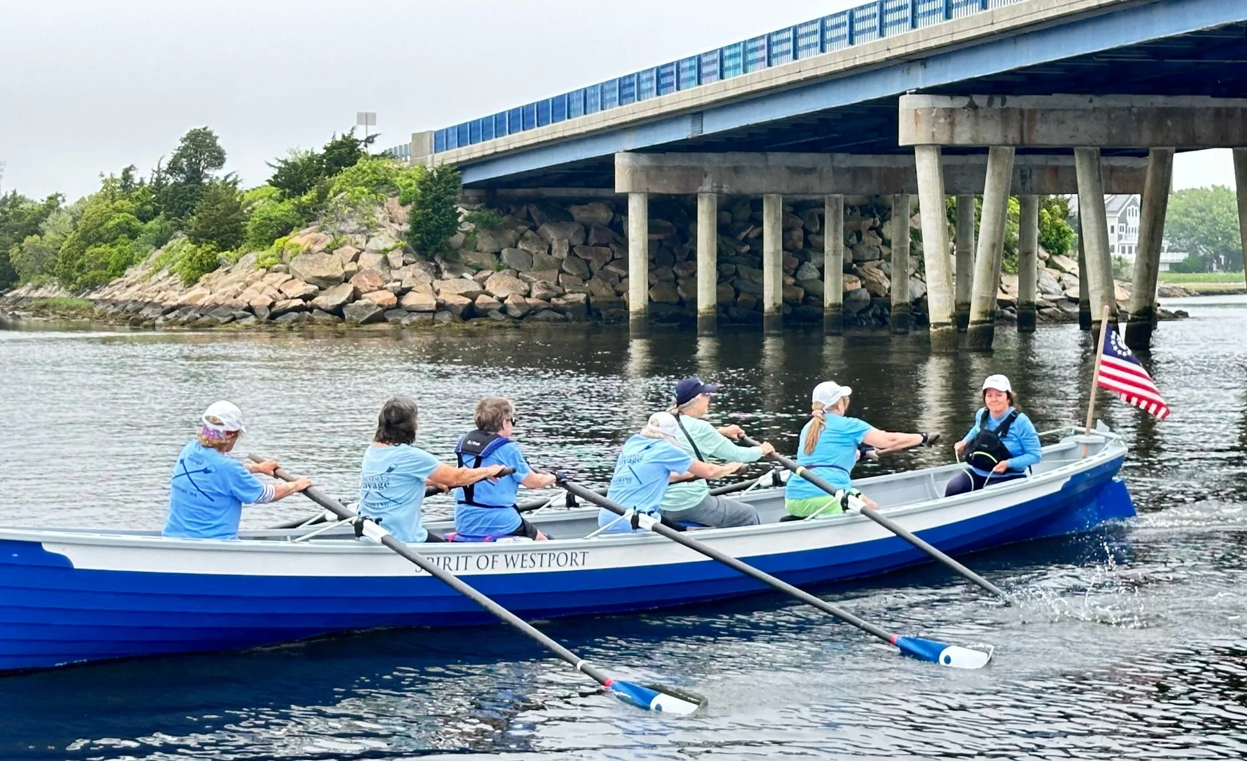 Women in a rowing boat on a river under a bridge, with greenery and rocks on the bank and an American flag at the stern.