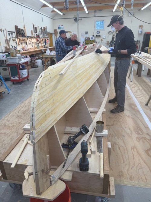 Wooden boat under construction in a workshop with tools and materials on workbenches, and drawings on the wall.