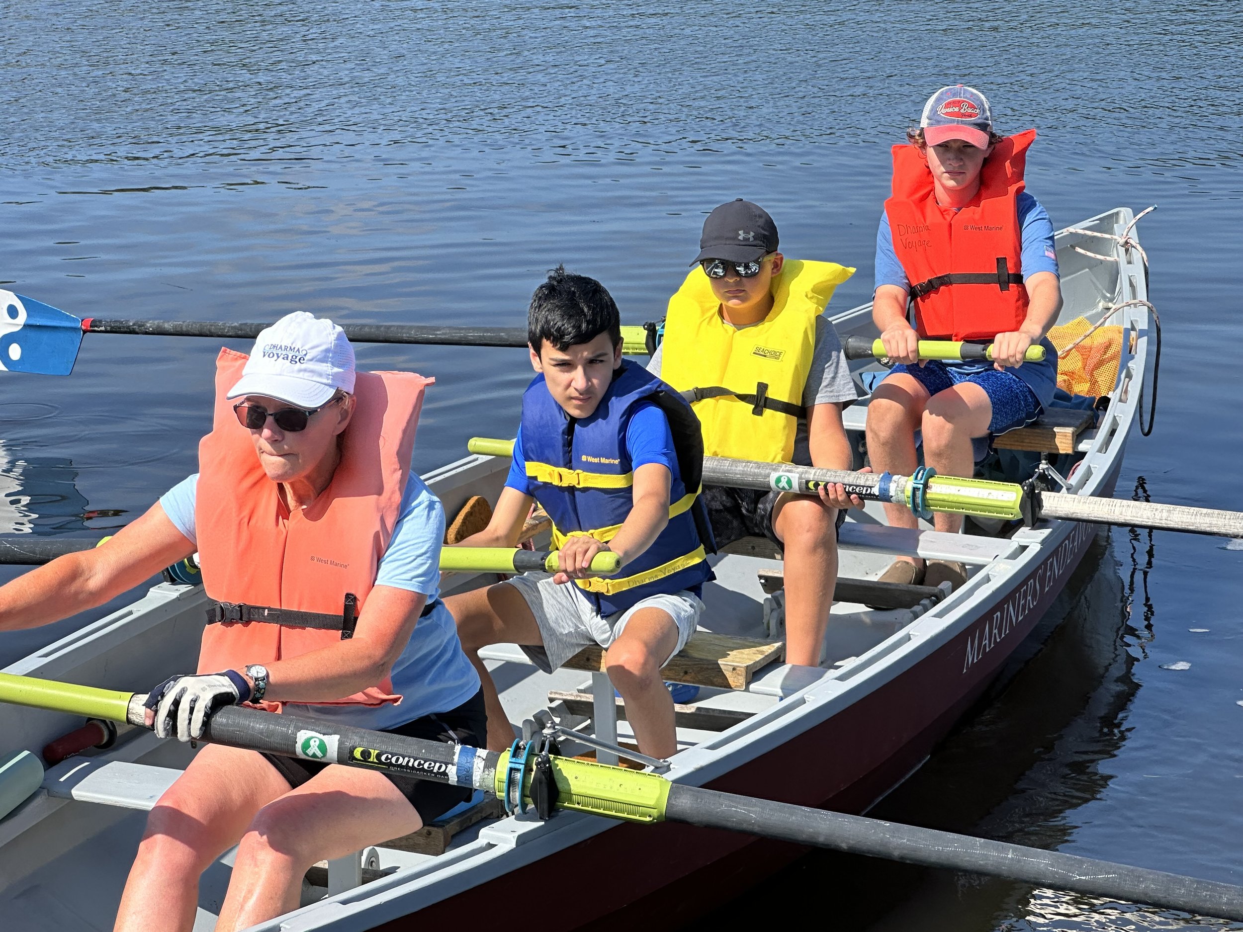Four children and an adult rowing a dory on the river, all wearing life jackets and hats.