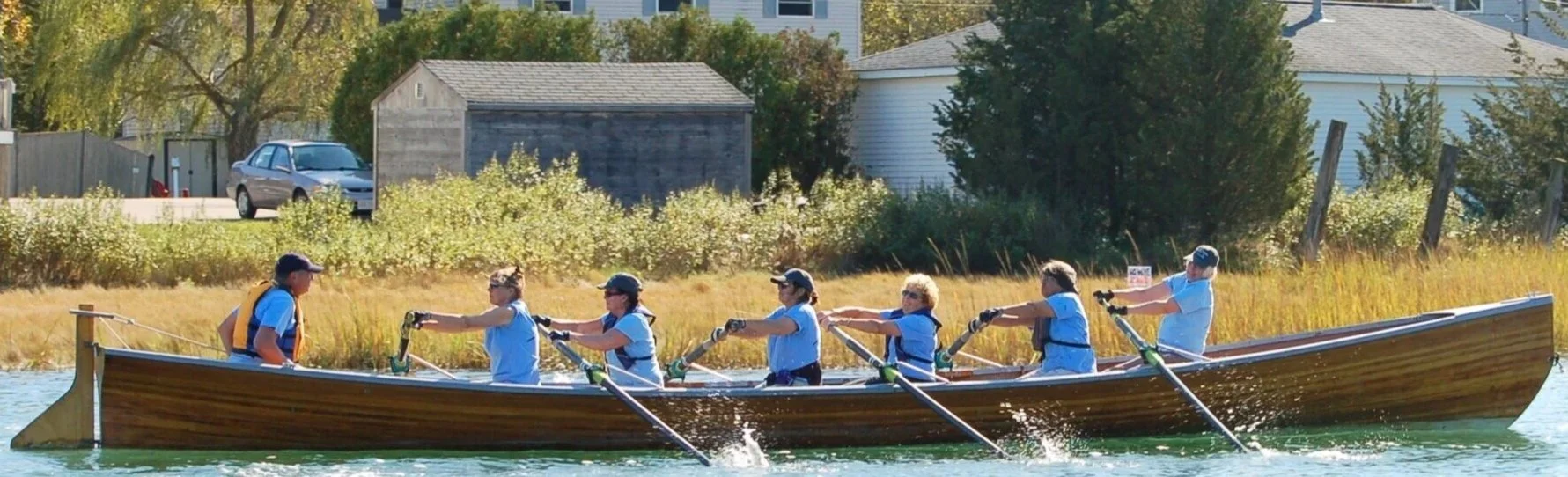 Group of women in a rowing boat on a river, under a bridge, with an American flag at the stern.
