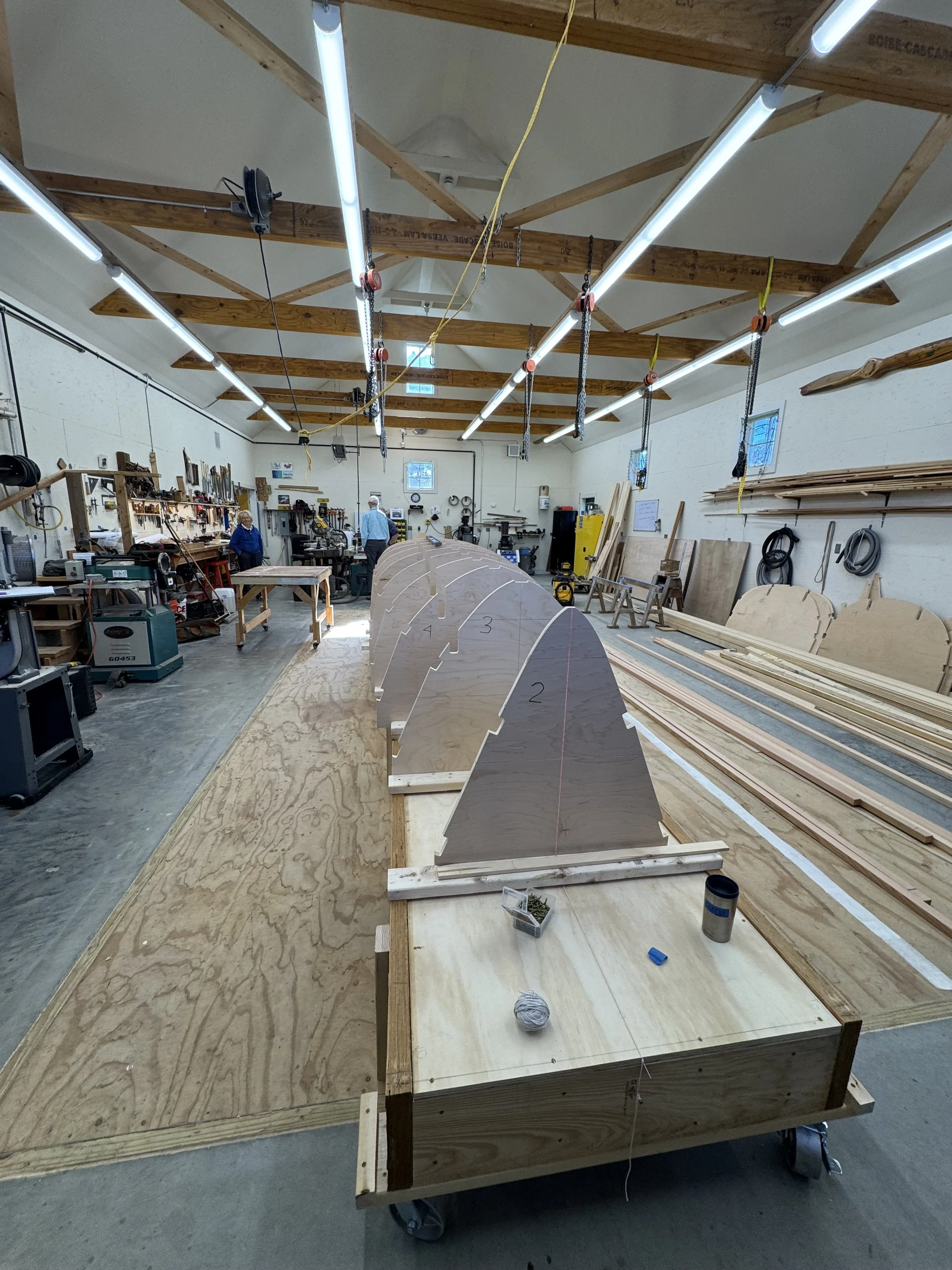 Wooden boat under construction in a workshop with tools, workbenches, and photographs on the walls.