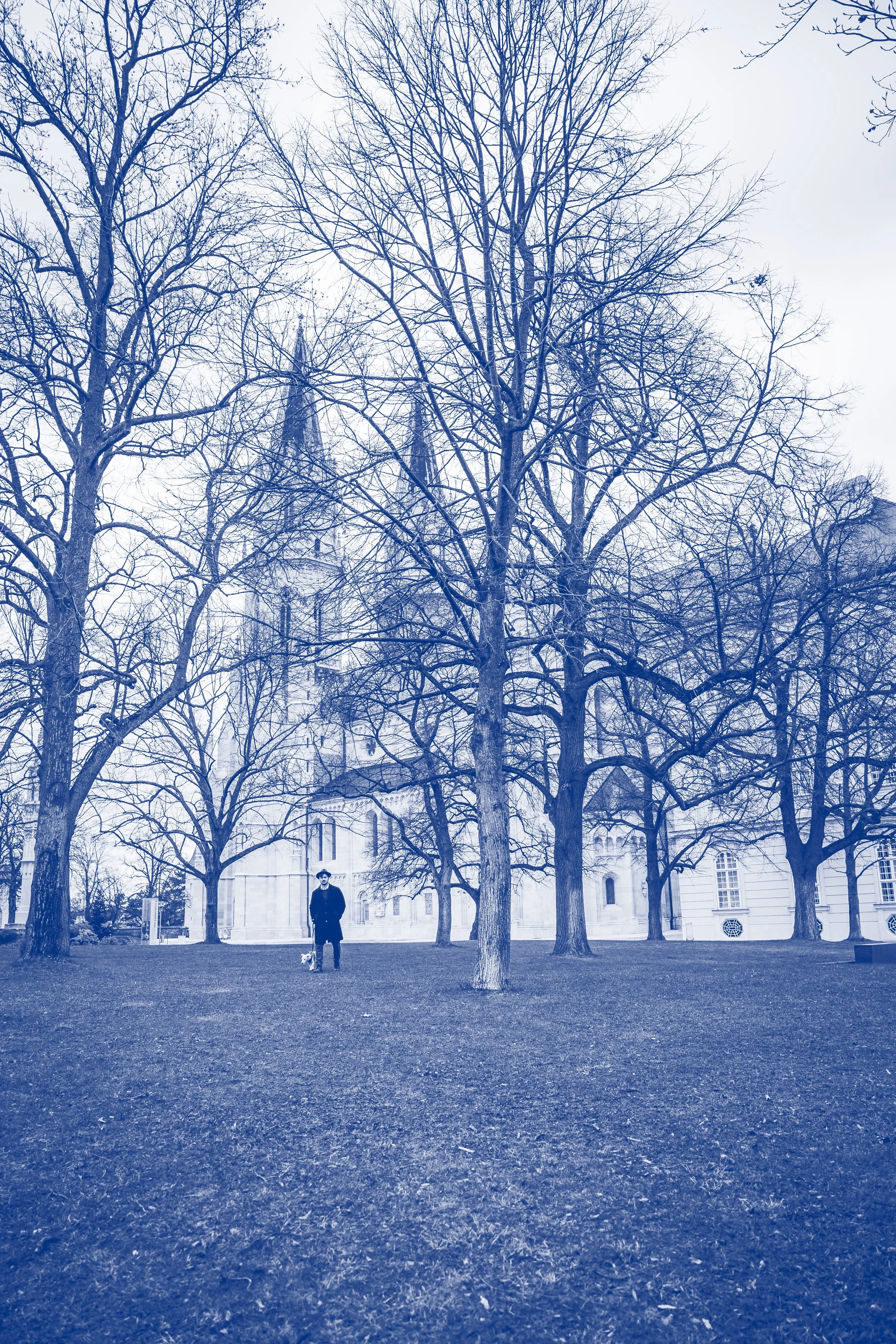 Ein Mann mit Hund steht in einem Park vor einer großen, alten Kirche. Es ist Winter, Bäume ohne Blätter umgeben die Kirche, der Himmel ist bewölkt.