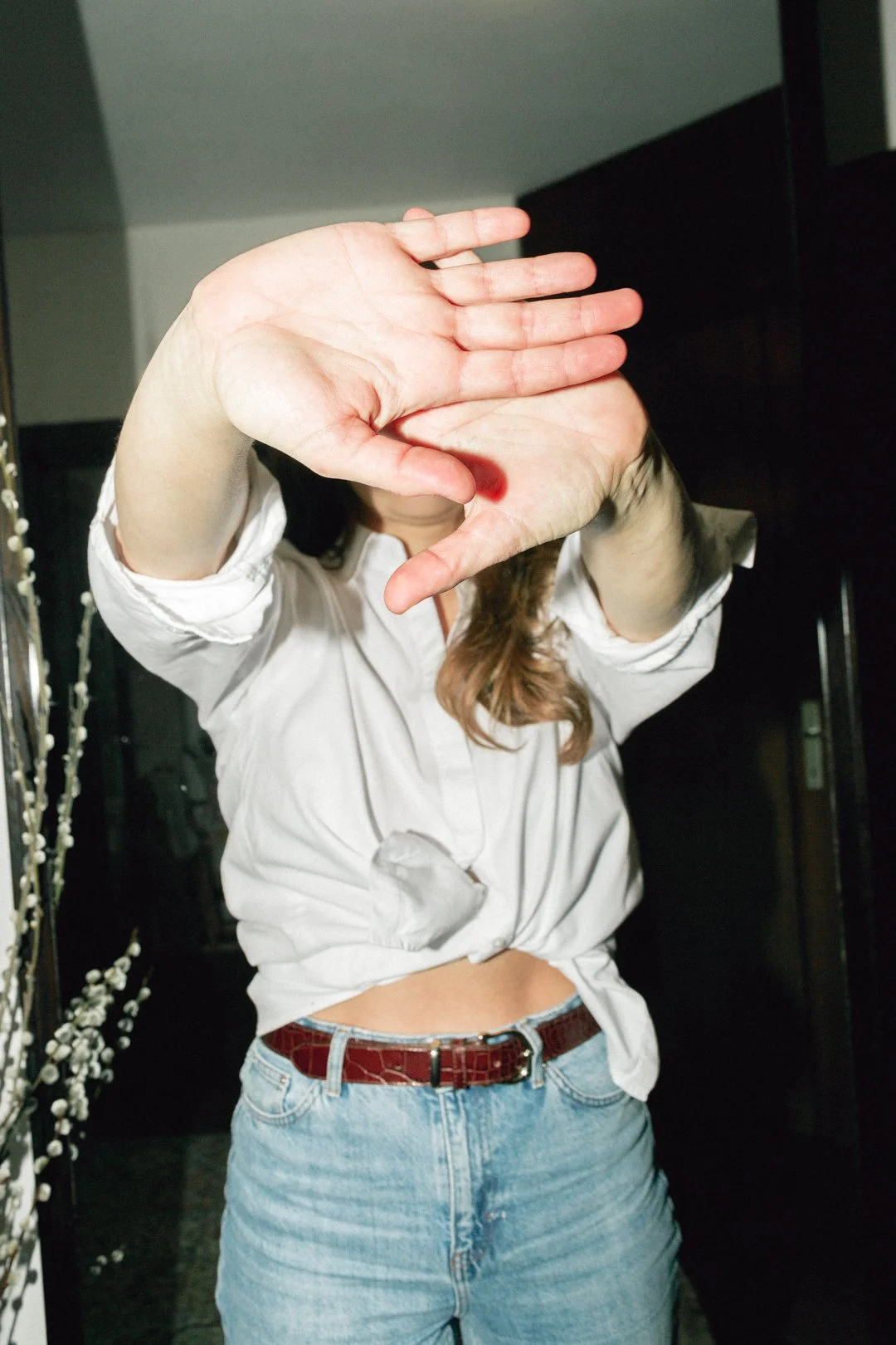 Frau mit weißen Hemd und Jeans macht eine Handgeste vor einer Kamera, ihr Gesicht ist hinter den Händen verborgen.