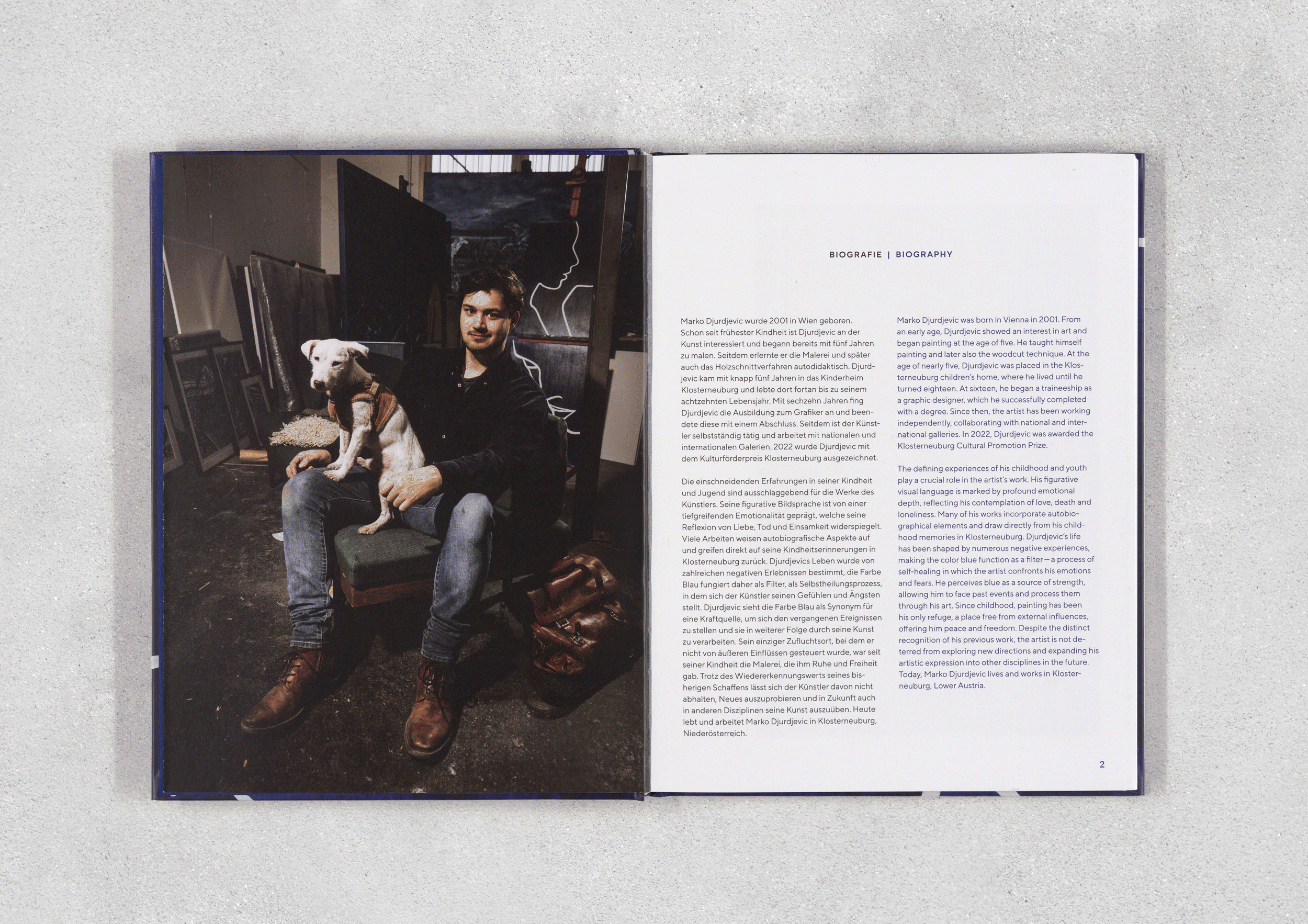 Offene Buch mit Text auf der rechten Seite, links Foto eines jungen Mannes mit Bart, dunklem Haar und gezeichneter Mütze, sitzt auf einem Stuhl, hält einen Welpen auf seinem Schoß, umgeben von Bildern und Kunstwerken im Hintergrund, im Raum mit dunklem Boden und grauer Wand.