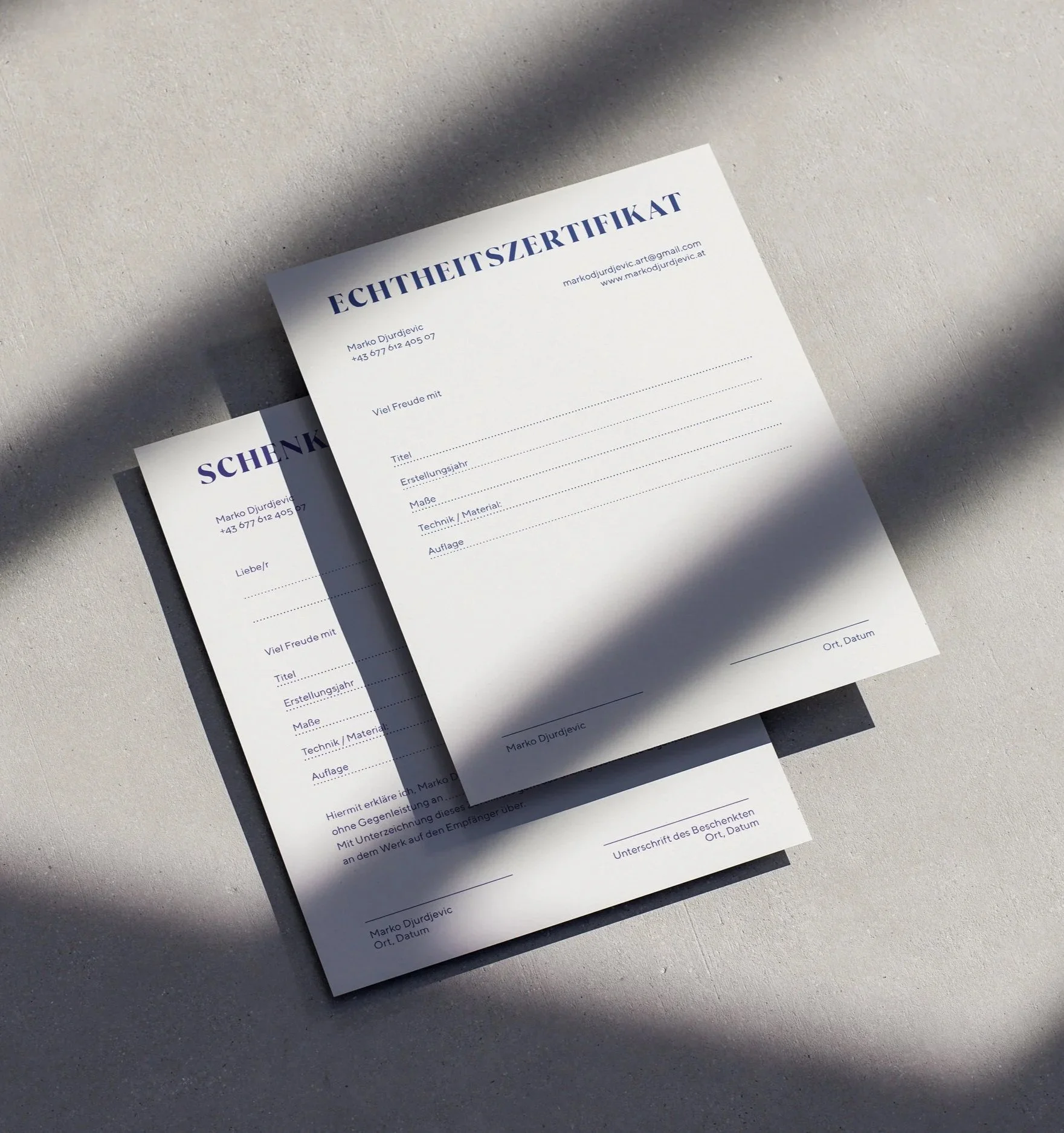 Zwei Dokumente, eins ist ein Zertifikat und das andere ein Geschenkpapier. Die Dokumente liegen auf einer hellen Oberfläche, mit Schatten von Blättern oder ähnlichem, die darauf fallen.