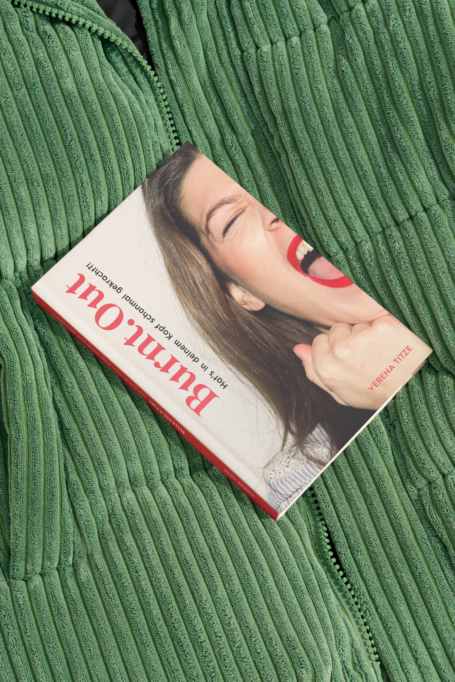 Ein Buch mit dem Titel "Bauchgefühl" von Verena Titze liegt auf einem grünen, gerippten Stoff, vermutlich einer Jacke. Das Buch zeigt ein lachendes Gesicht einer Frau mit rote Lippen.