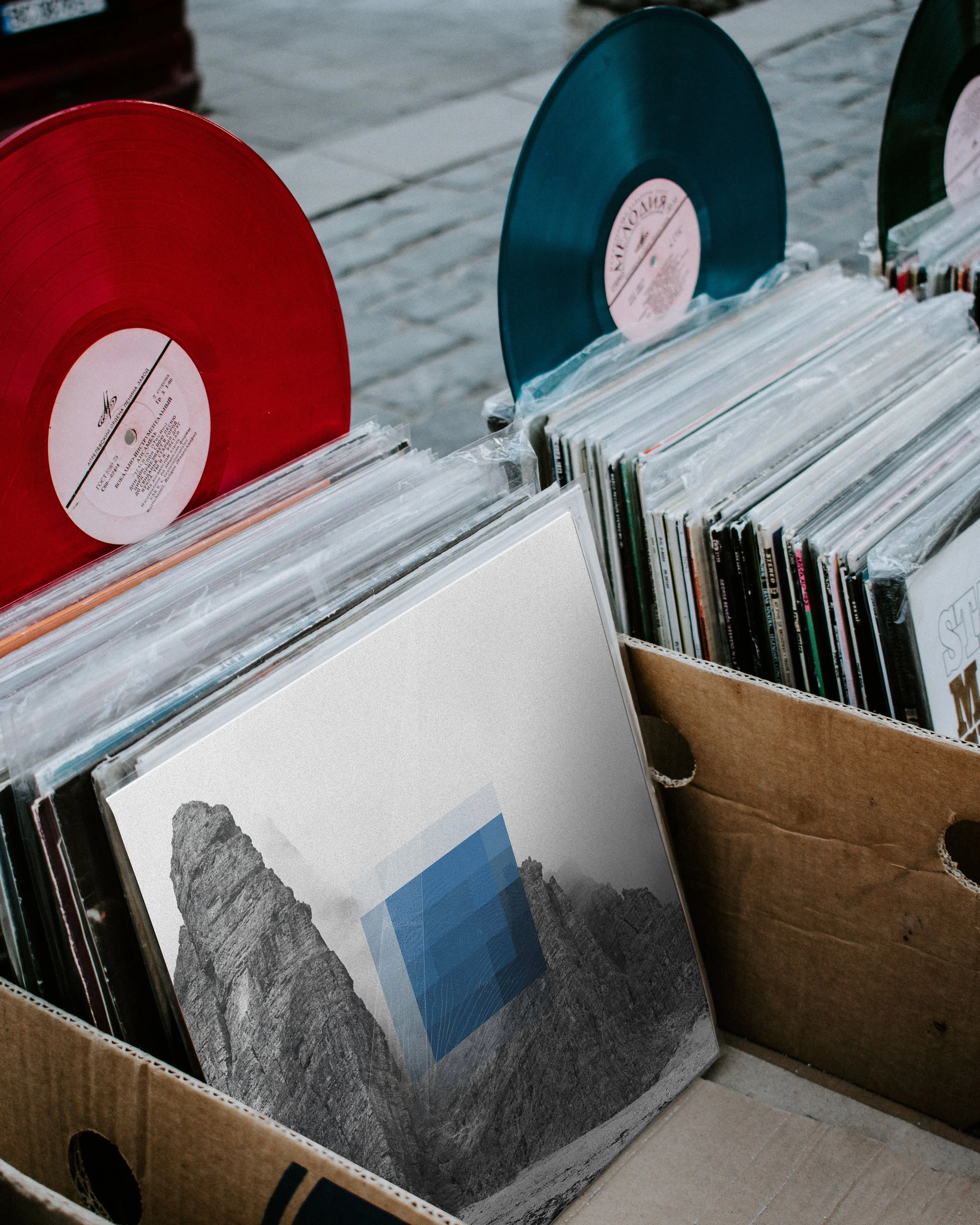 Vinyl-Schallplatten liegen in einem Karton auf einem Marktständer, einige sind in Plastikfolie verpackt, darunter eine mit einem schwarzen und weißen Cover mit einem Berg-Design, auf dem eine blaue 3D-Quadrat-Form abgebildet ist.