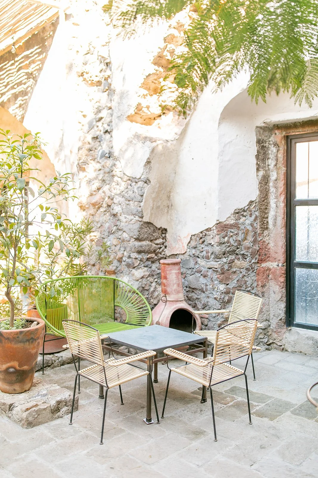 An outdoor patio featuring a table, four chairs, potted plants, a rustic chimney, and a weathered stone wall with a large window, surrounded by greenery.
