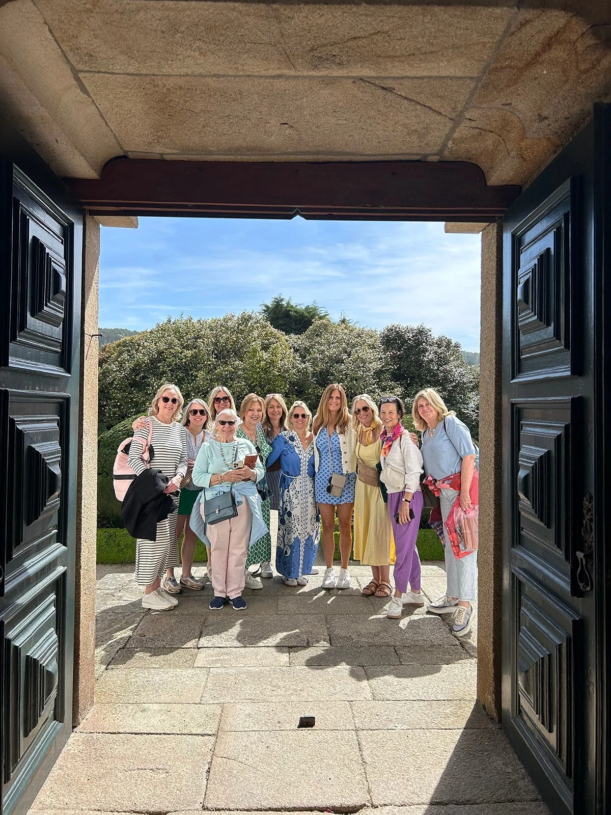 A group of women standing outdoors near a garden, viewed through an open stone doorway, with a blue sky and trees in the background.