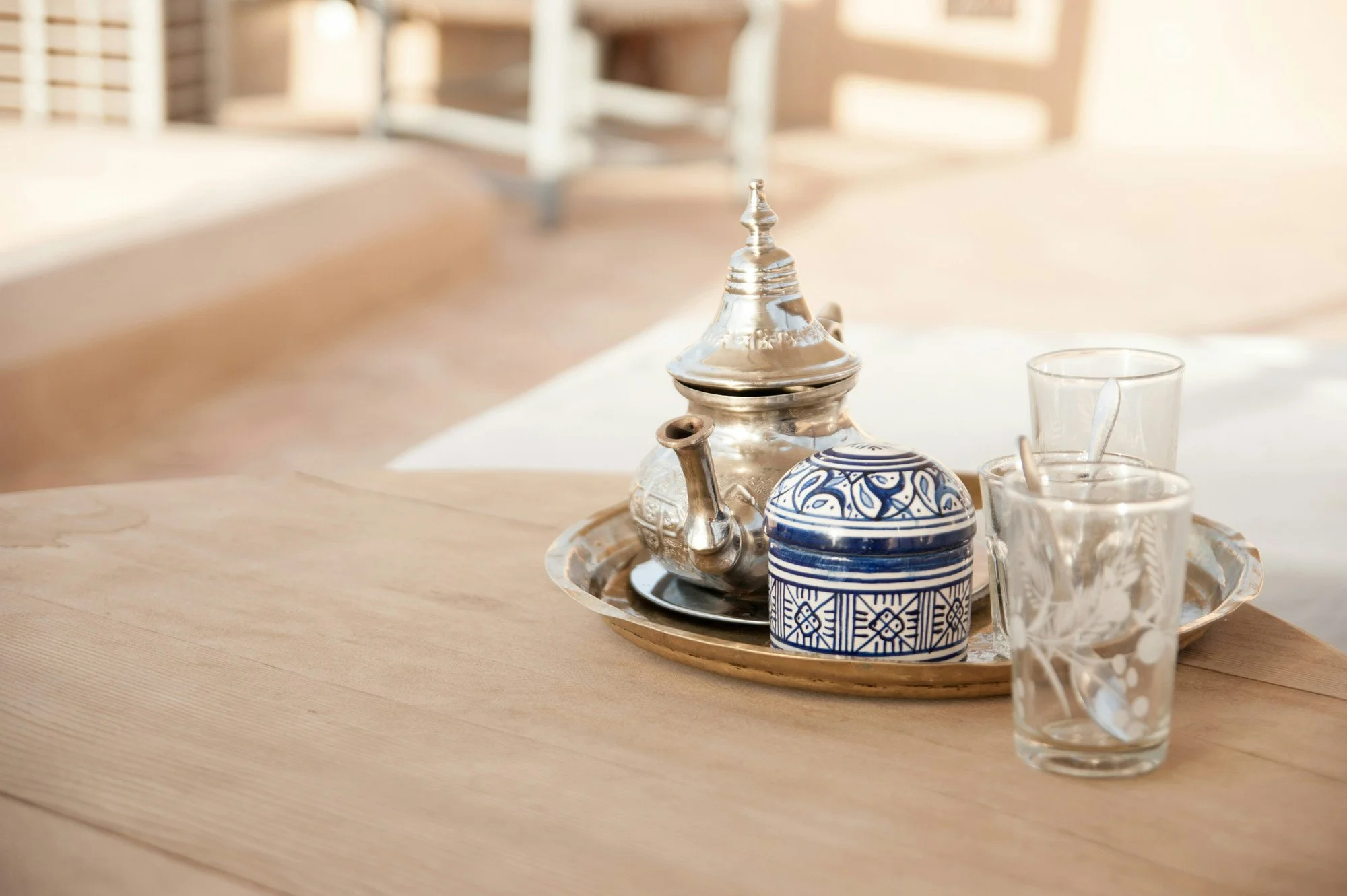 A wooden table with a decorative metal tray holding a silver teapot and a blue and white patterned container, flanked by two clear glasses with spoons inside, in a well-lit room with wooden chairs in the background.