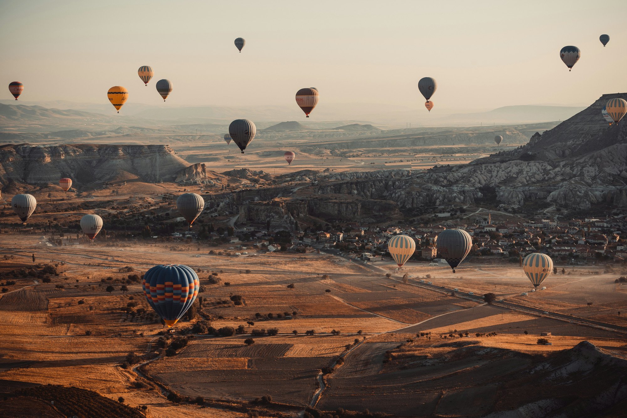 Multiple hot air balloons floating over a landscape with rocky formations, fields, and a small town at sunrise or sunset.