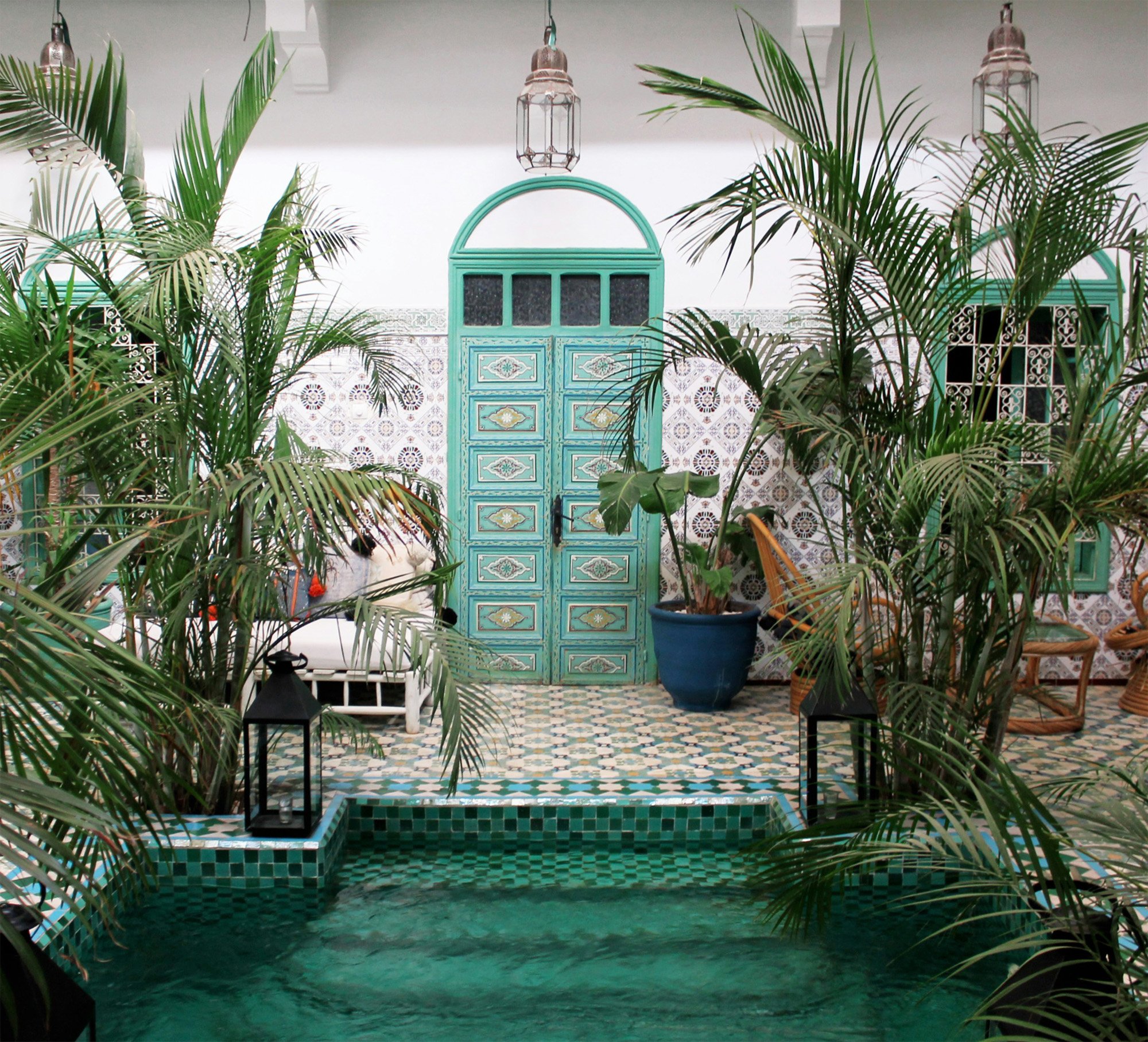 Indoor courtyard with Moroccan-style decor, featuring a small pool surrounded by green plants, ornate turquoise door, patterned tile floor, and wicker chairs.