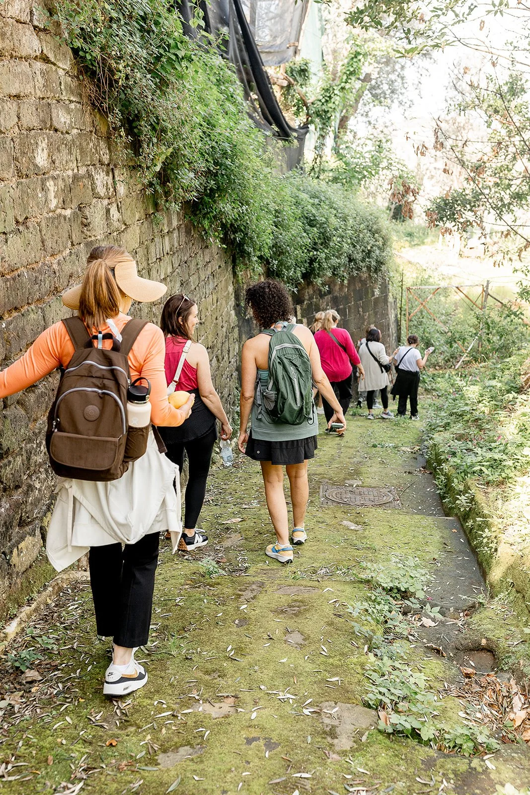 A group of women hiking along a moss-covered trail beside a brick wall, surrounded by lush green foliage and trees.