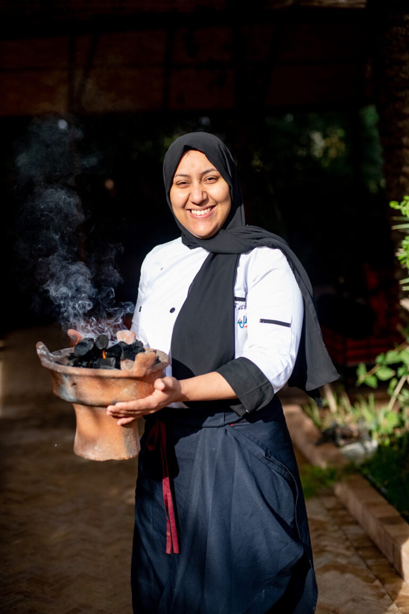 A woman wearing a black hijab and a white chef's coat holding a clay pot with glowing charcoal and smoke.