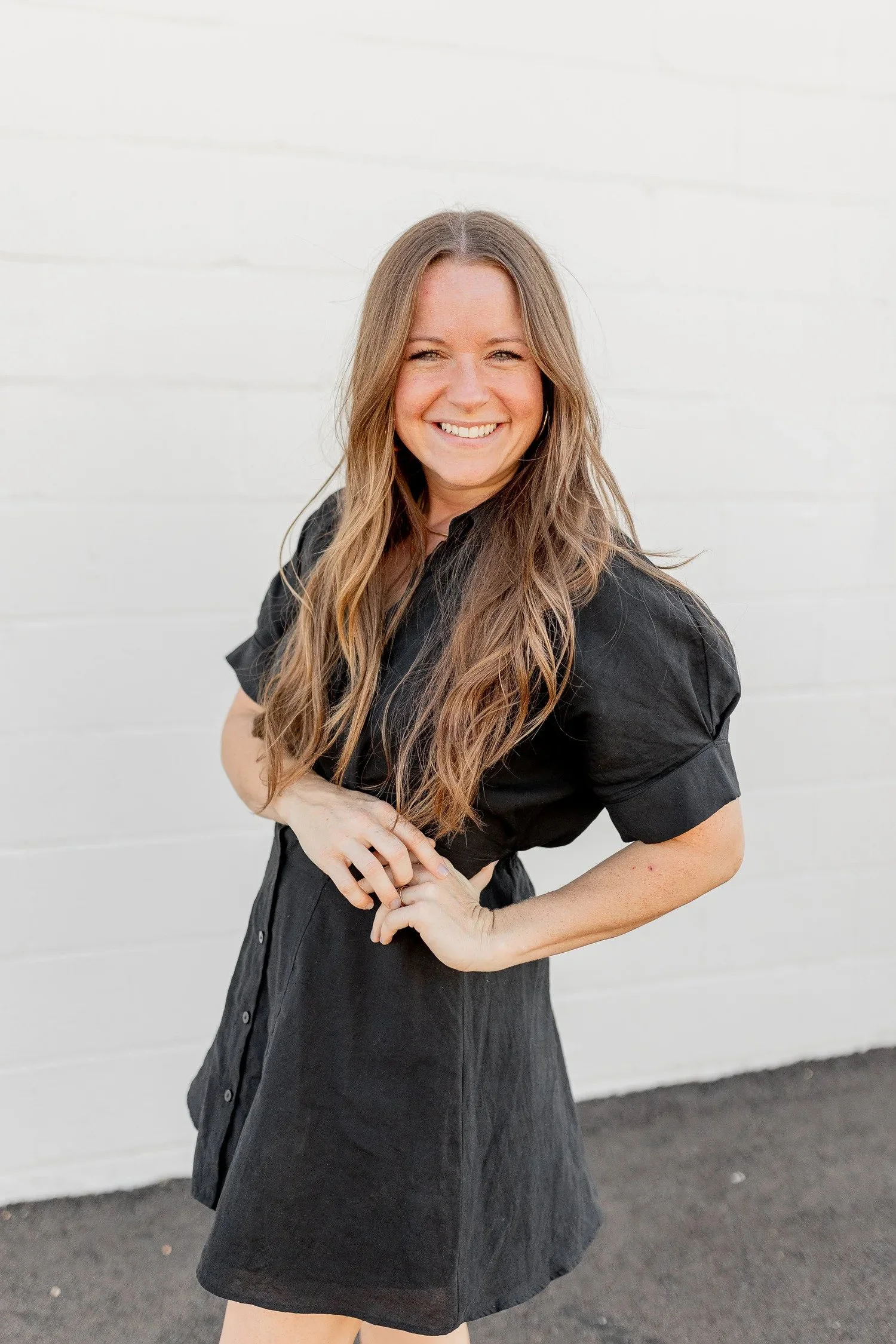 A woman with long wavy brown hair, wearing a black short-sleeve dress, smiling and posing against a white brick wall.