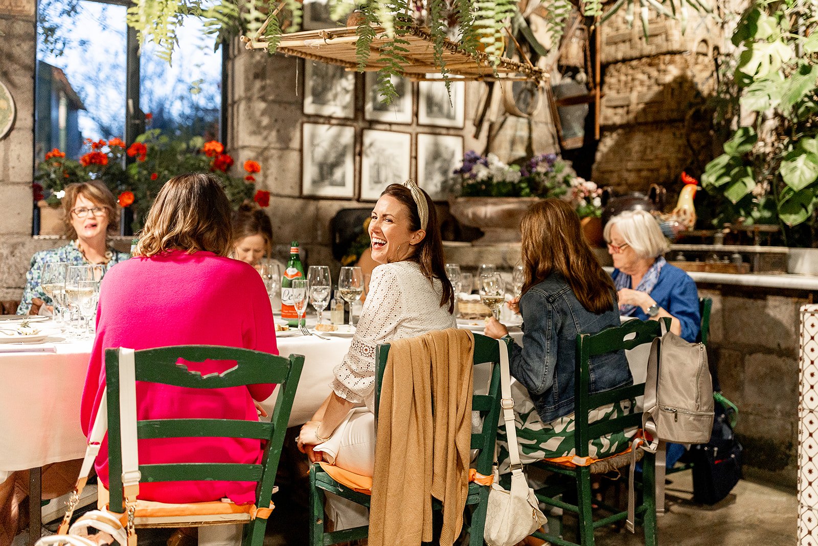 A group of women sitting at a dining table, enjoying a meal and conversation in a rustic setting with plants and flowers.