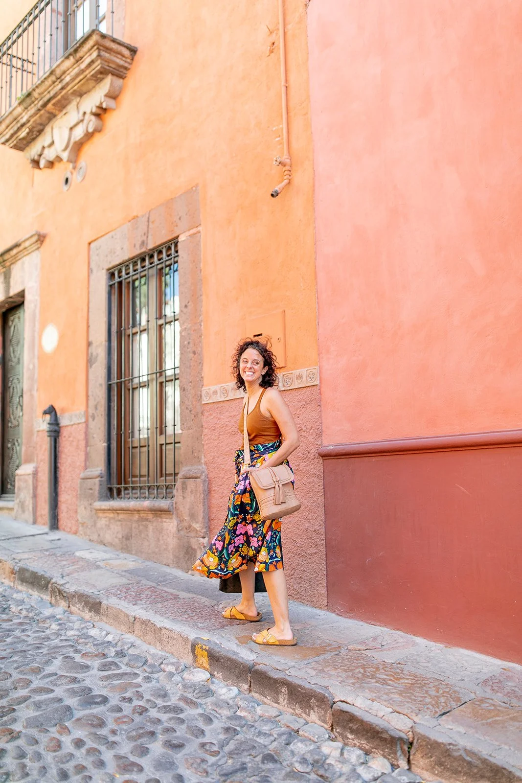 A woman with curly hair wearing a brown top, a colorful floral skirt, and yellow sandals, smiling and standing on a cobblestone street against an orange building with barred windows.