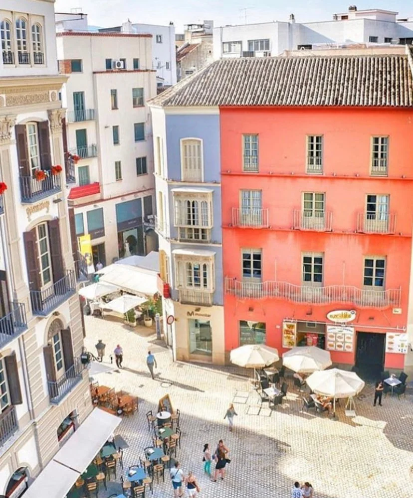 A European city square with colorful historic buildings, outdoor tables with umbrellas, and pedestrians walking.