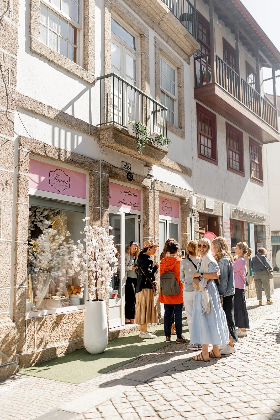 A group of people standing outside a bakery or cafe with pink signs, on a cobblestone street in front of a stone building with balconies and flower pots.