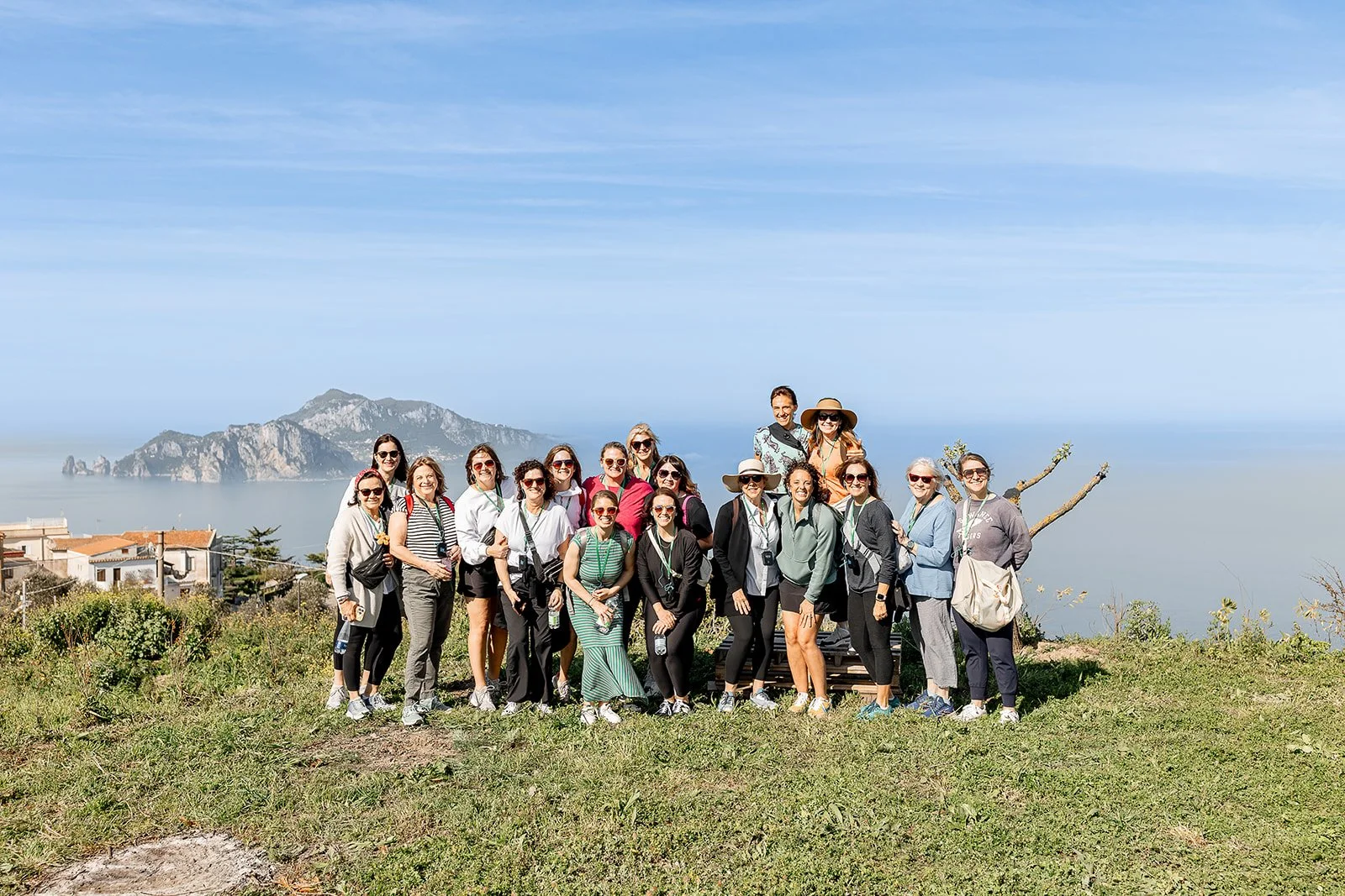 Group of women and two men posing outdoors with a mountainous coastline in the background, on a grassy hill under a blue sky.