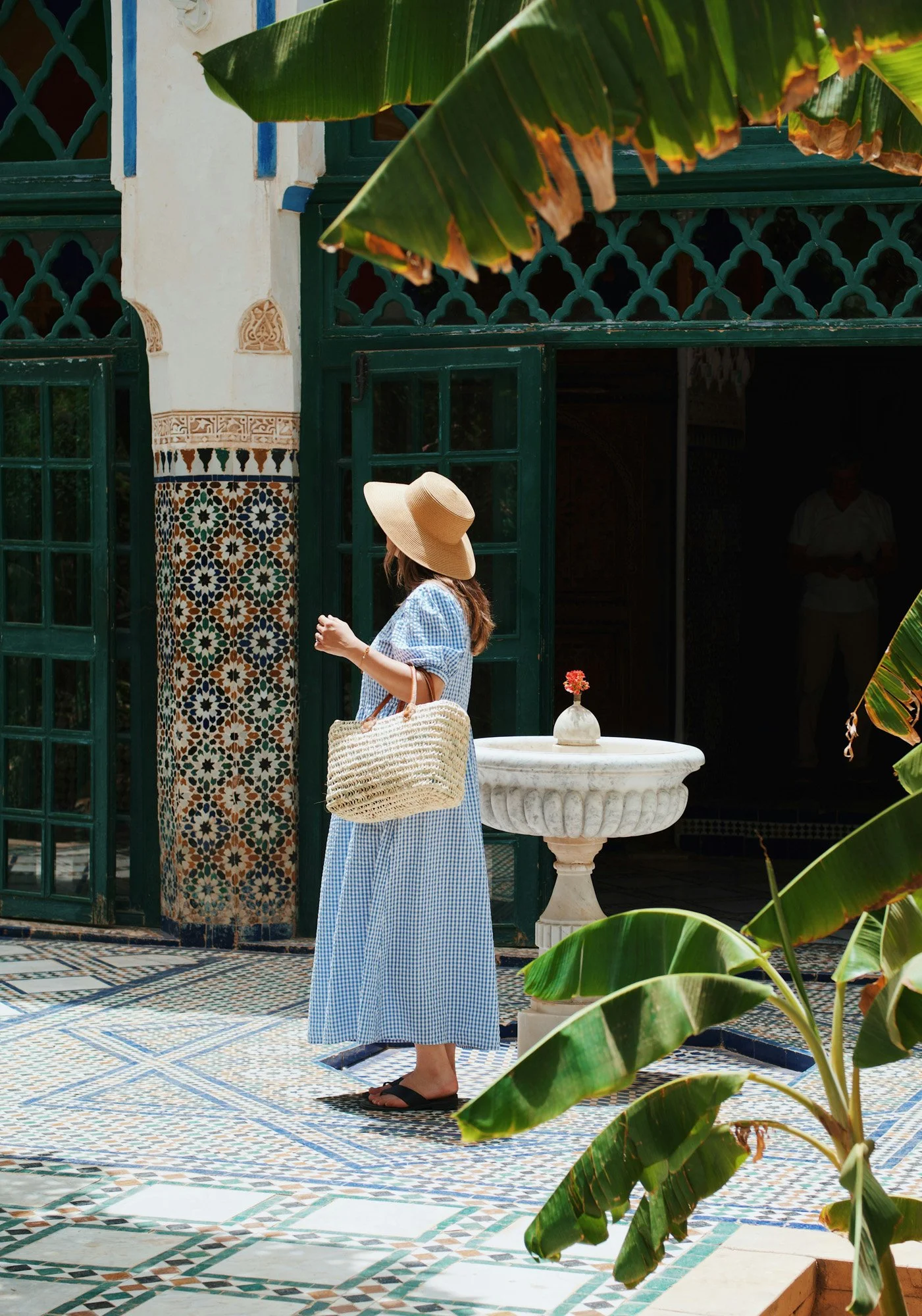 A woman in a blue checkered dress, wearing a wide-brimmed straw hat and black sandals, stands next to a white stone fountain with a small flower in a vase on it. She carries a woven straw bag and is outside a building with green doors and intricate tilework, surrounded by large green leaves.