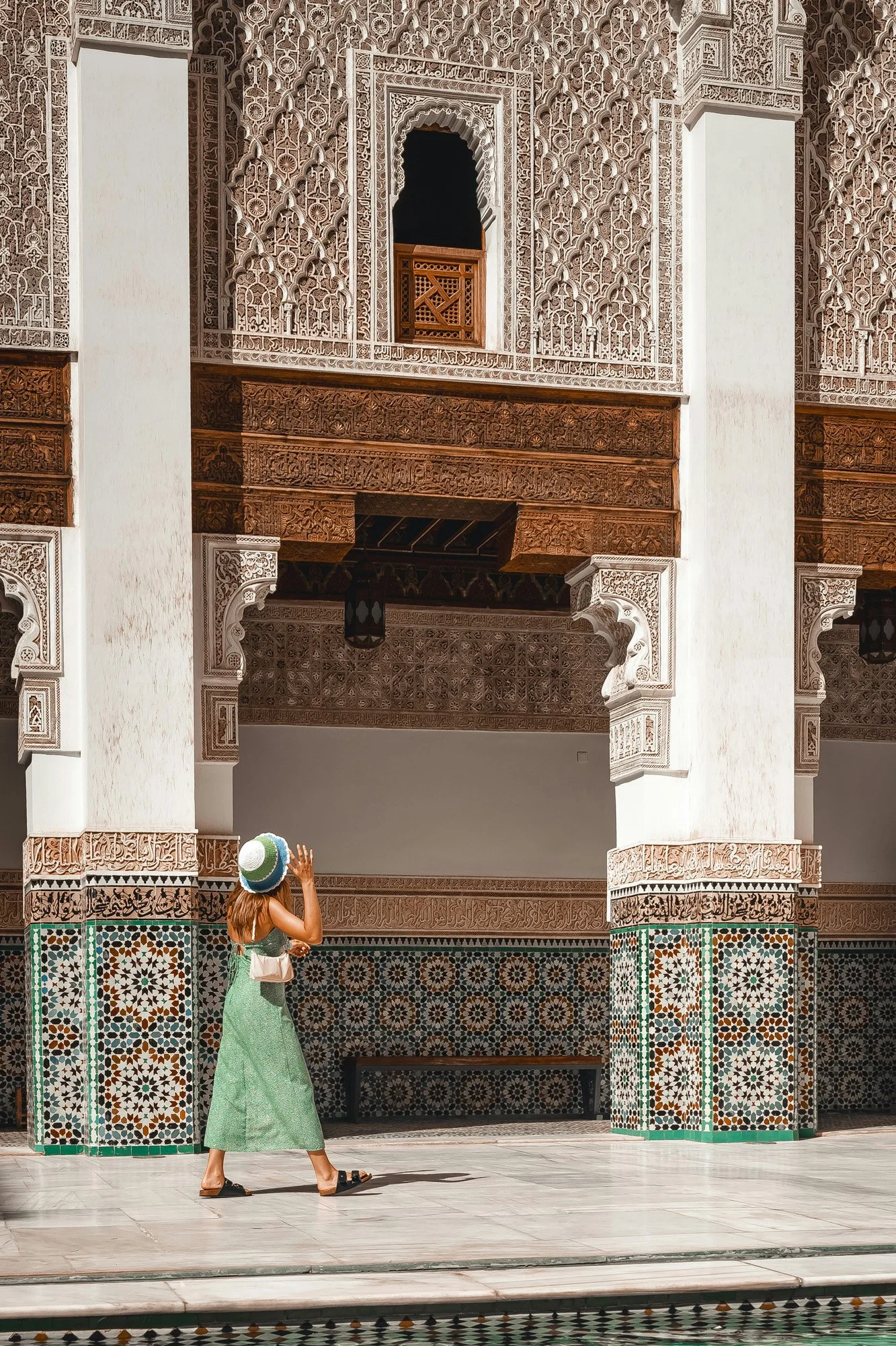 A woman in a green dress and large sun hat walking in front of ornate architectural walls with intricate Moroccan-style tile work.
