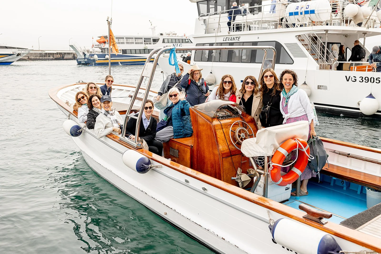 A group of women enjoying a boat ride on a harbor, smiling and posing for the camera. The boat has a wooden steering wheel and a lifebuoy at the stern.