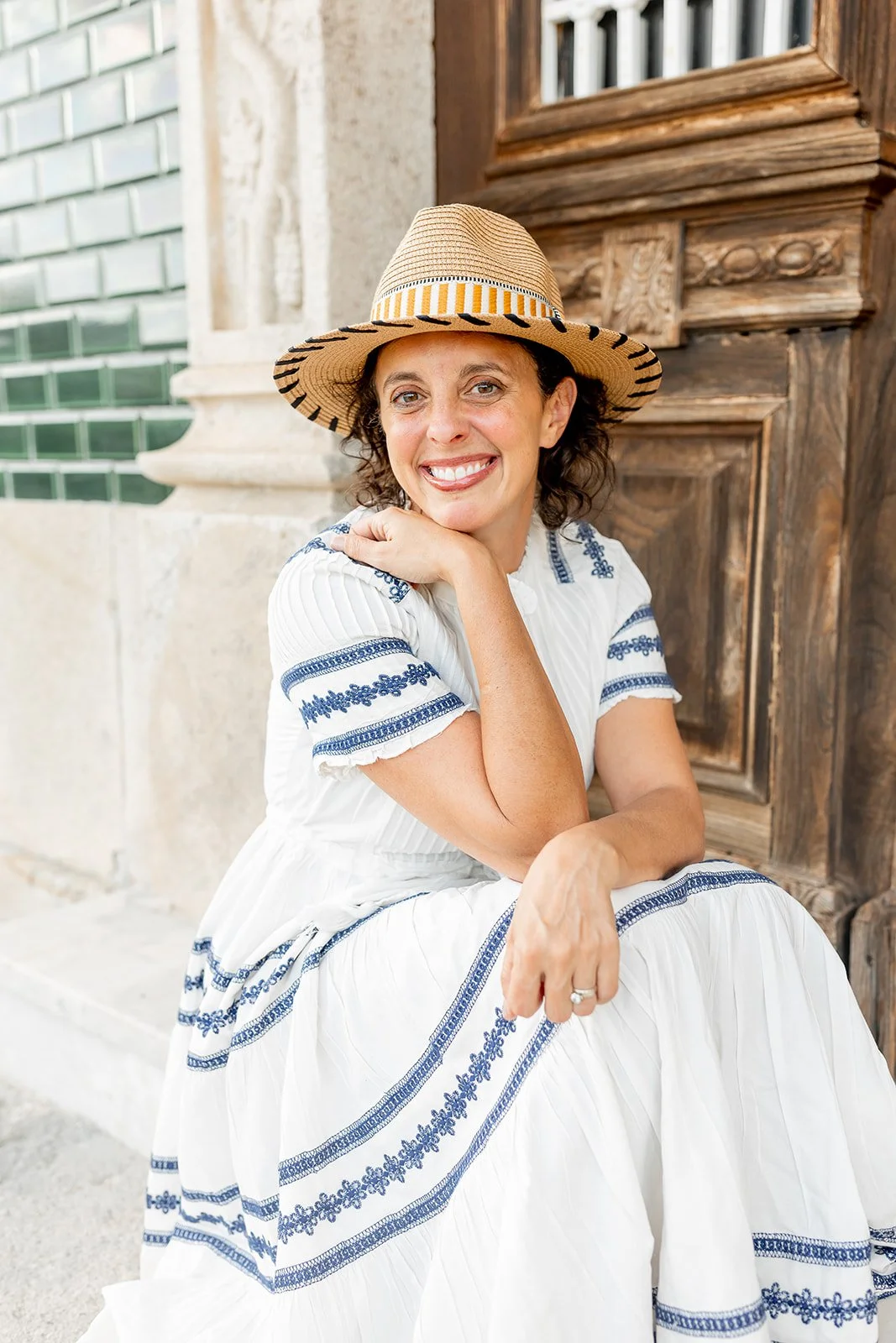 Woman in a white dress with blue embroidery, wearing a wide-brimmed straw hat, sitting beside a wooden door outside.