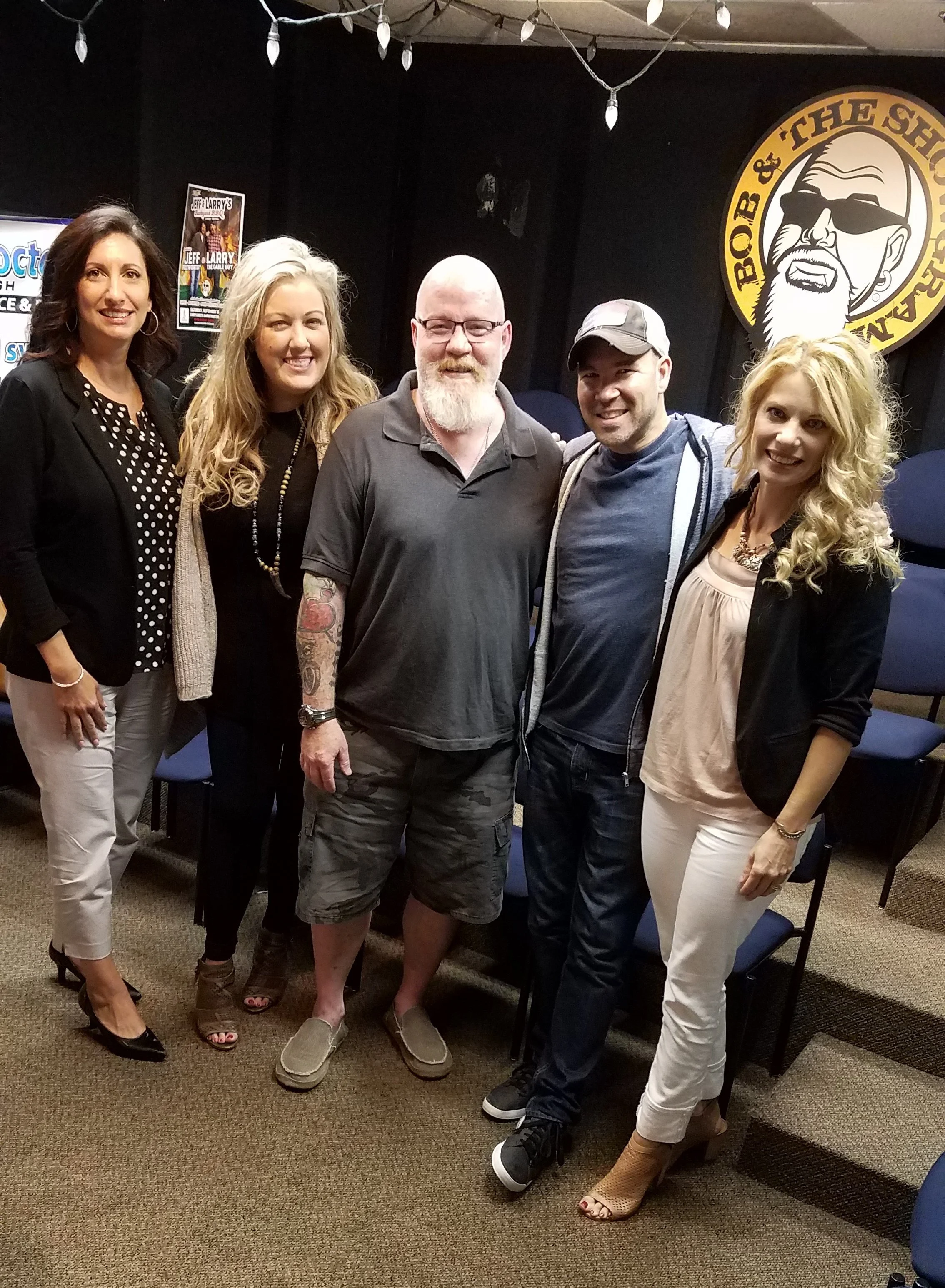 Group of five people standing together and smiling in a room with a black wall. There is a logo with a bearded man near sunglasses in the background. The group includes three women and two men, dressed in casual and semi-formal attire.