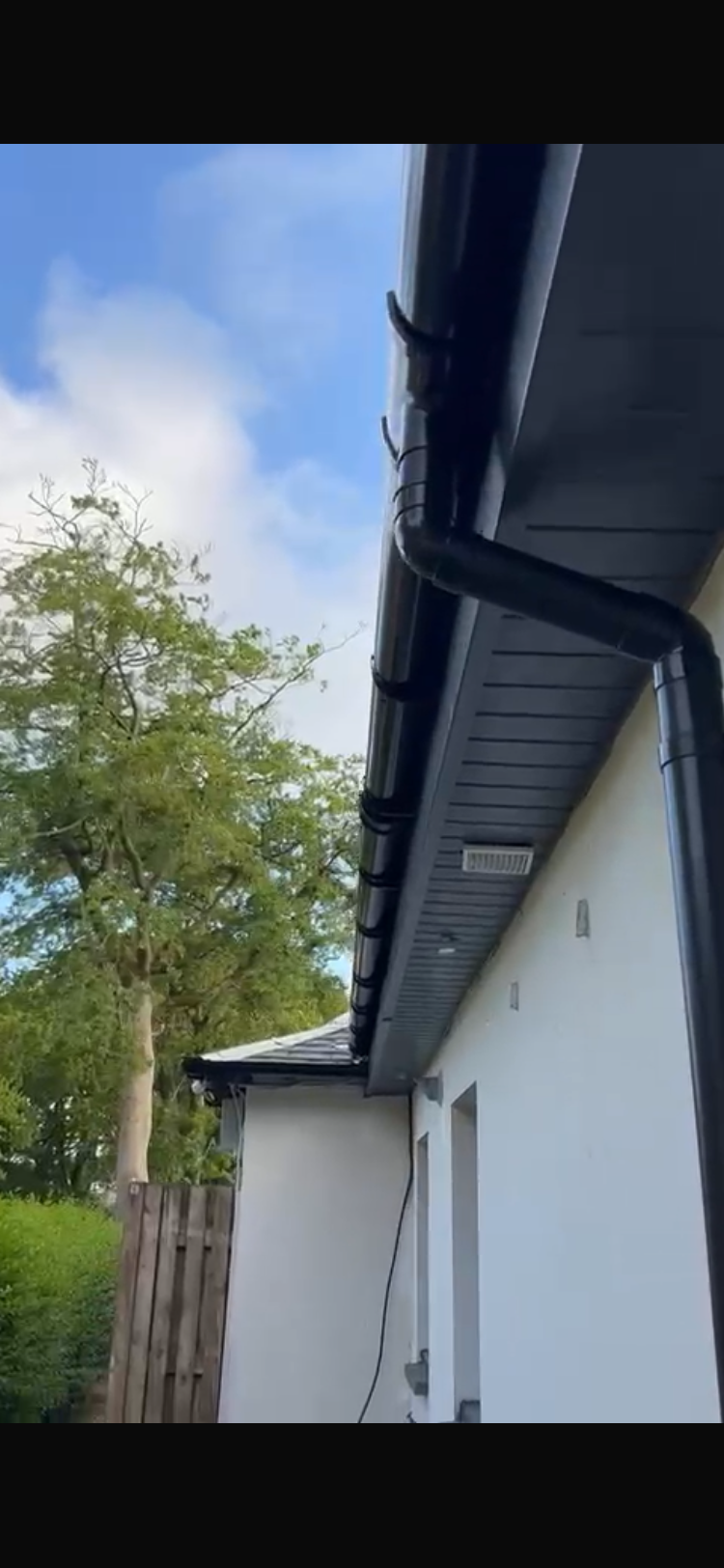 A close-up view of a house's gutter system with black rain gutters and downspouts, with a tree and cloudy sky in the background.