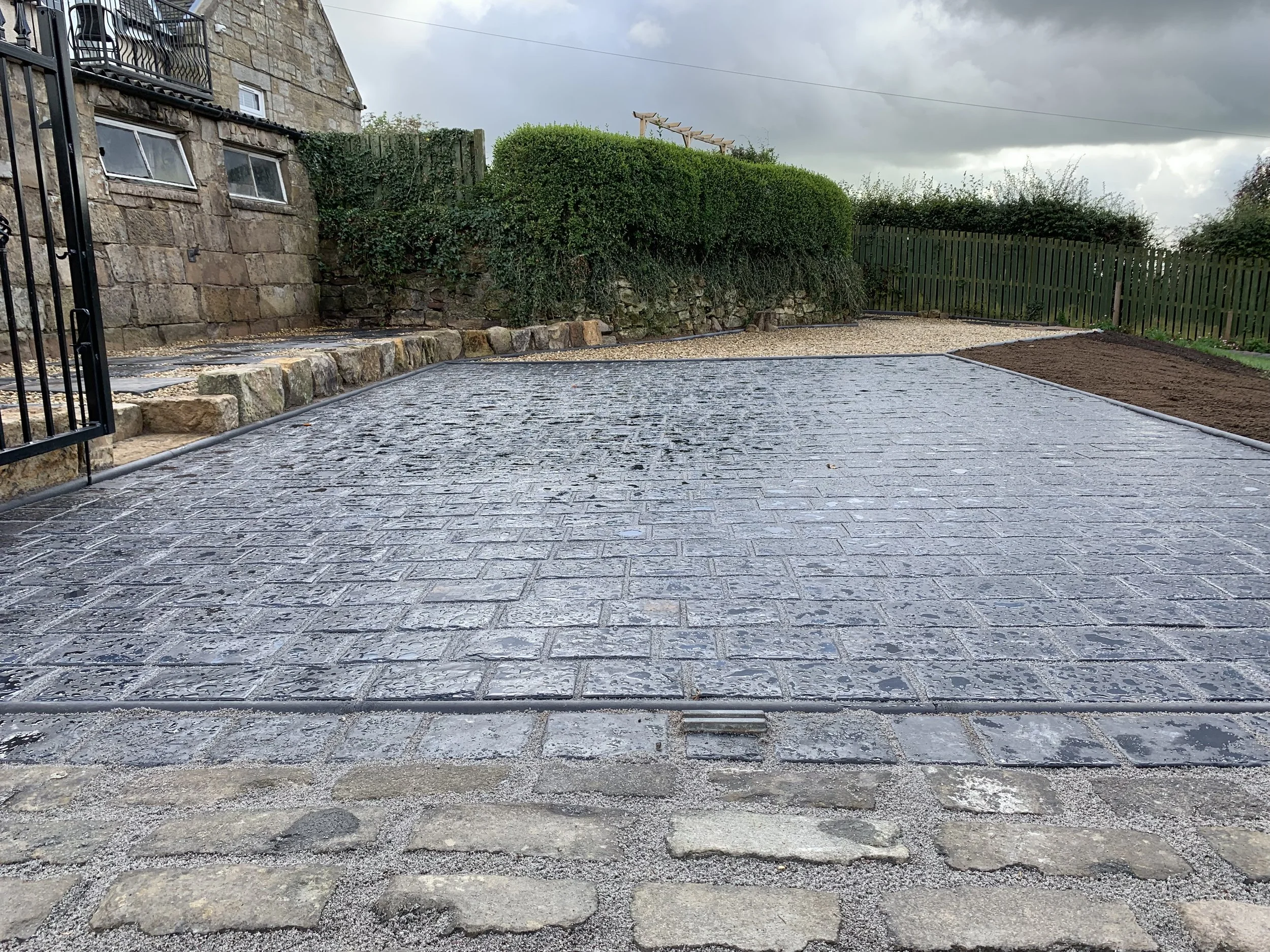 A freshly paved brick pathway in a backyard, with a stone curb at the front and a metal gate on the left side. In the background, there are green bushes, a stone retaining wall, a wooden fence, and an overcast sky.