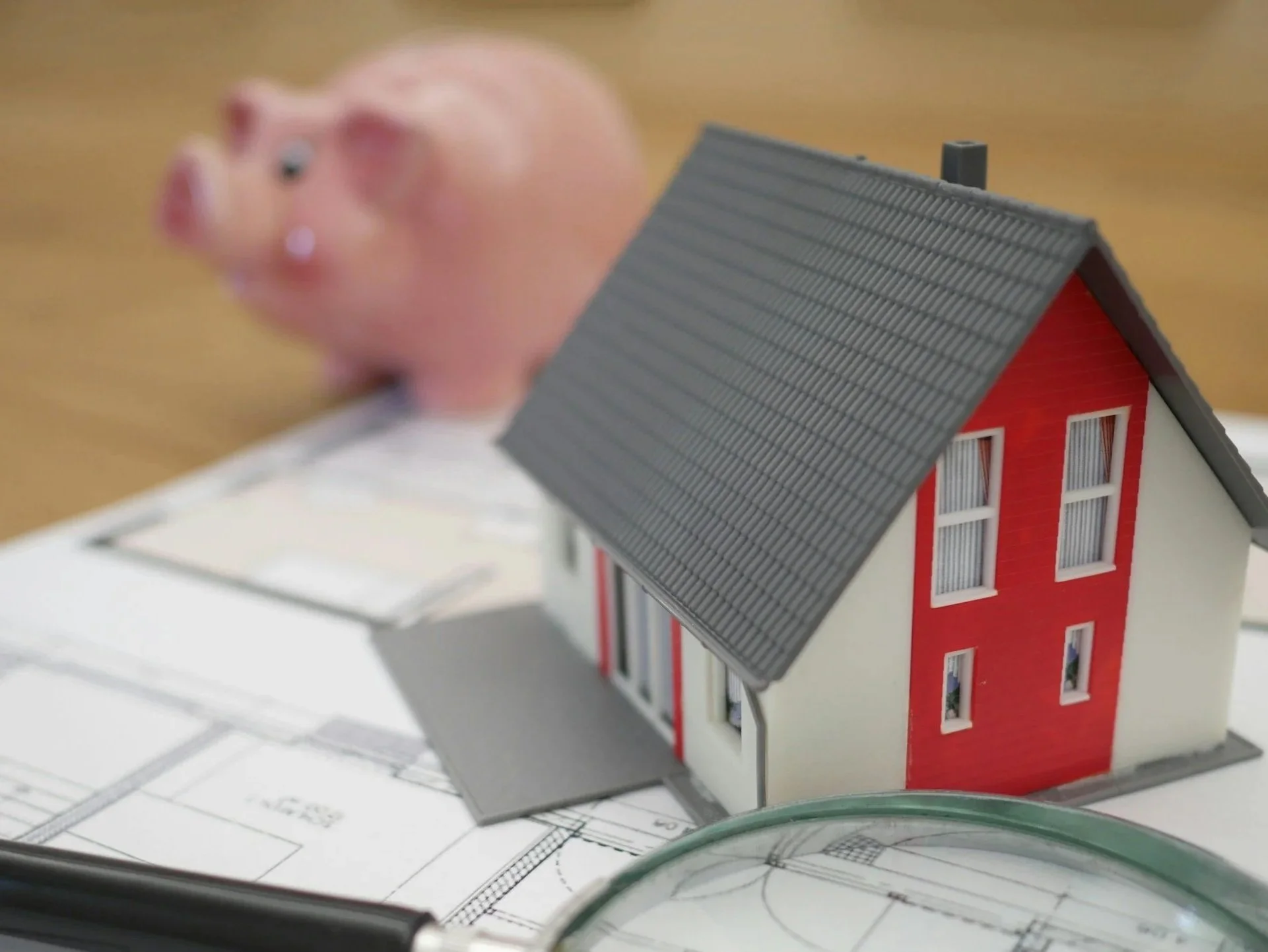 A miniature model of a house placed on architectural blueprints with a piggy bank in the background.