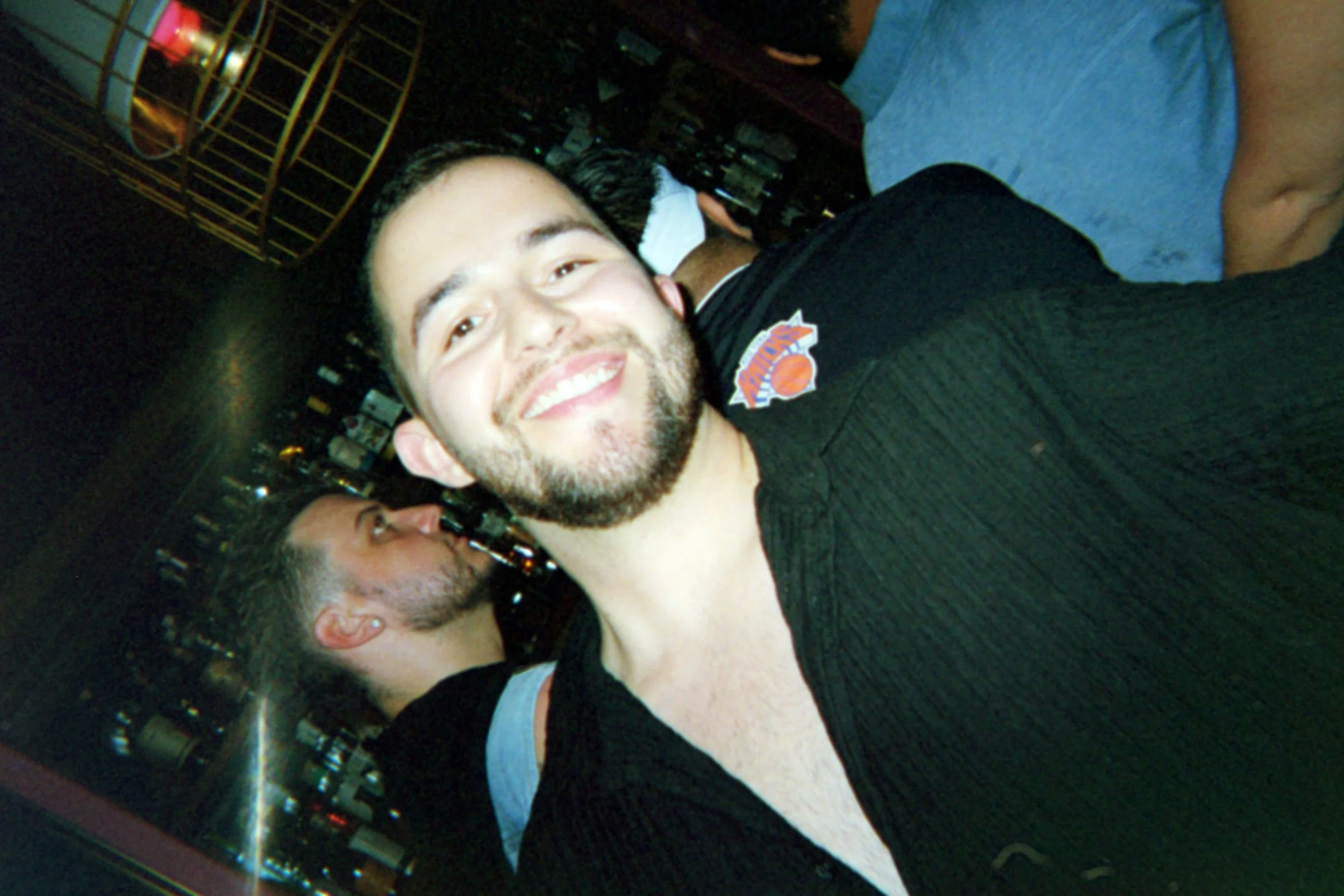 A smiling man with a beard taking a selfie in a bar, with a woman kissing him on the cheek in the background.