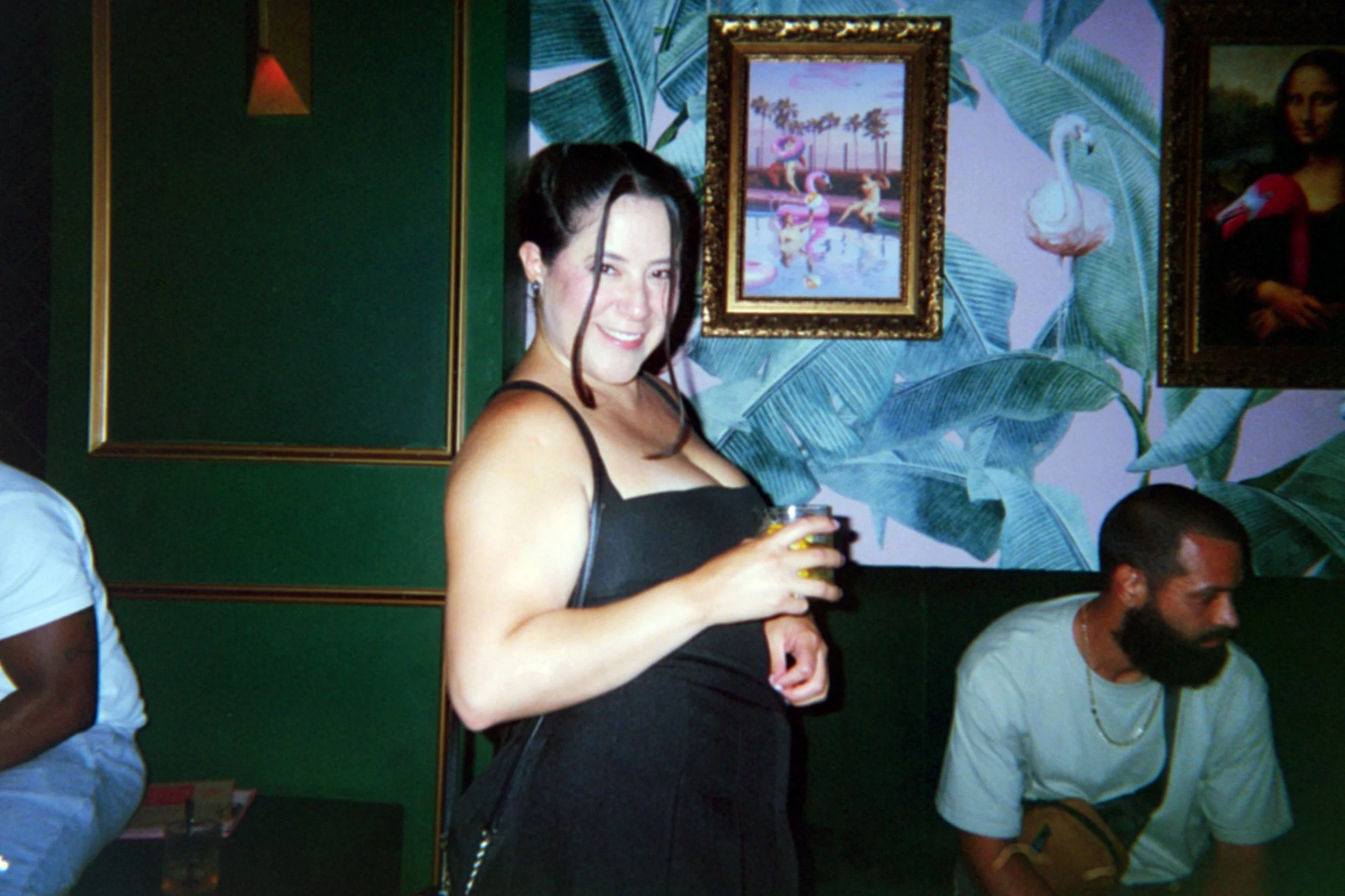 A woman with dark hair smiling and holding a drink, standing in a room with tropical-themed wallpaper and framed artwork, while a man with a beard sits nearby.