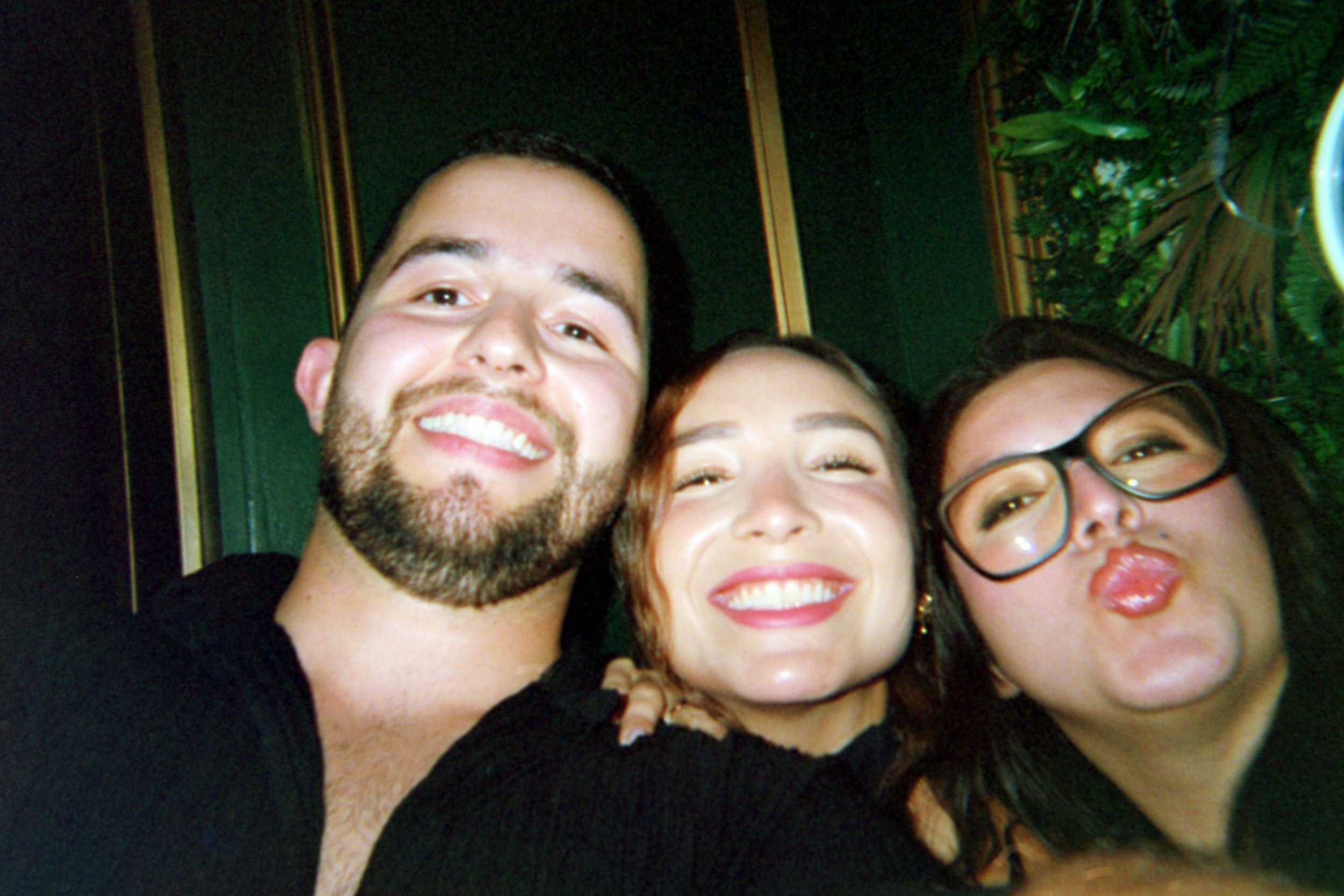 Three friends taking a selfie together, smiling, with a green background and some plants visible.