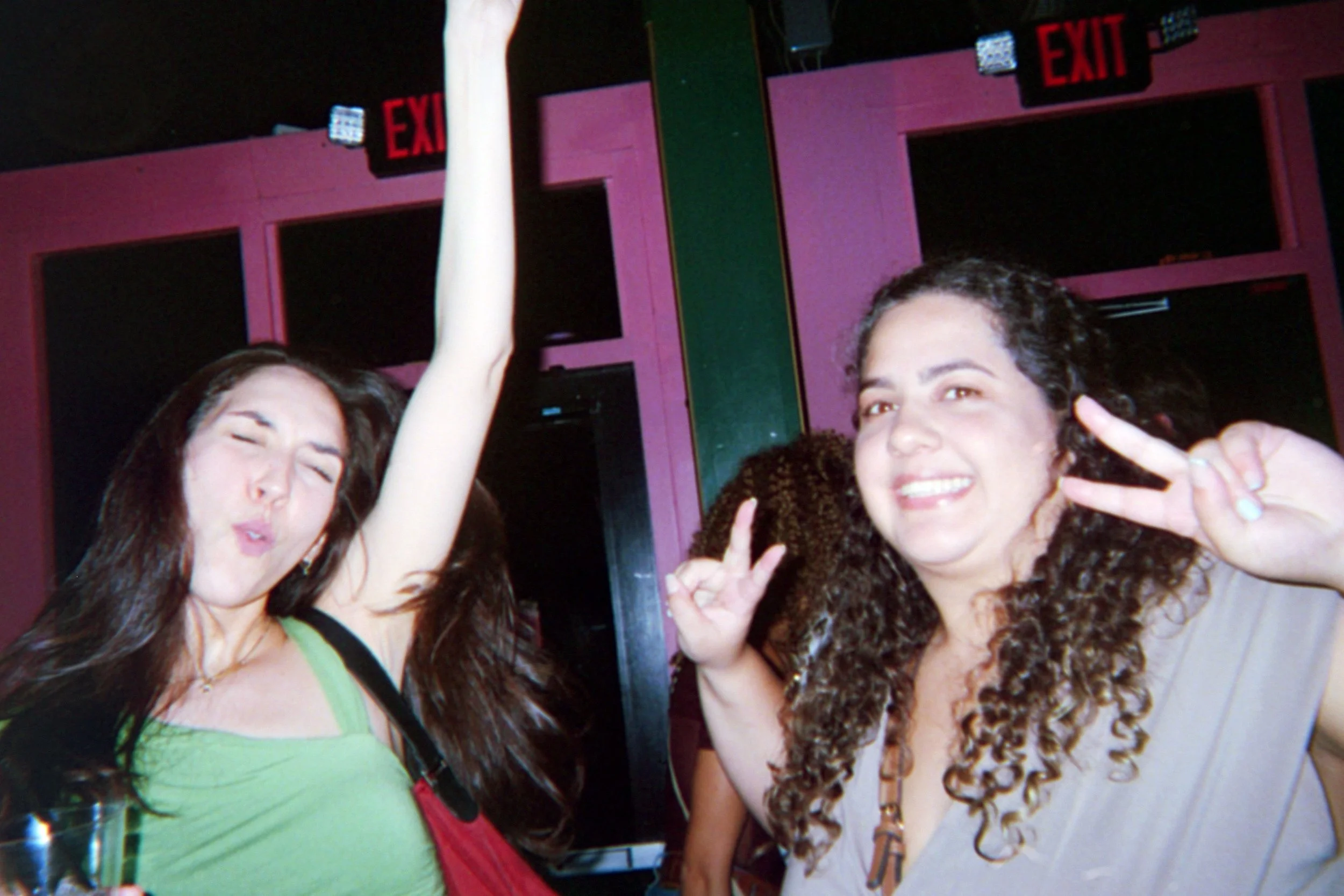 Two women dancing and smiling at a party with pink walls and red exit signs in the background.