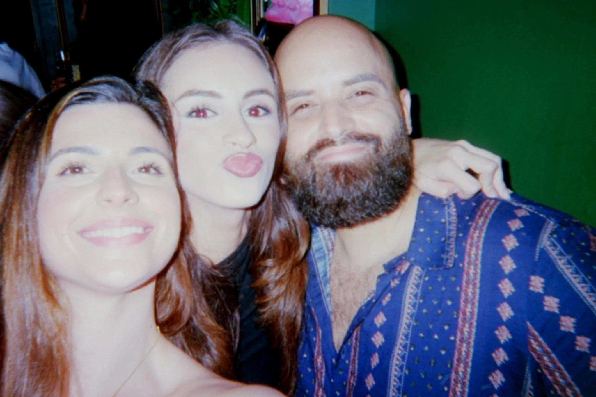 Group of three friends taking a selfie together, smiling and making playful expressions, indoors against a green wall.
