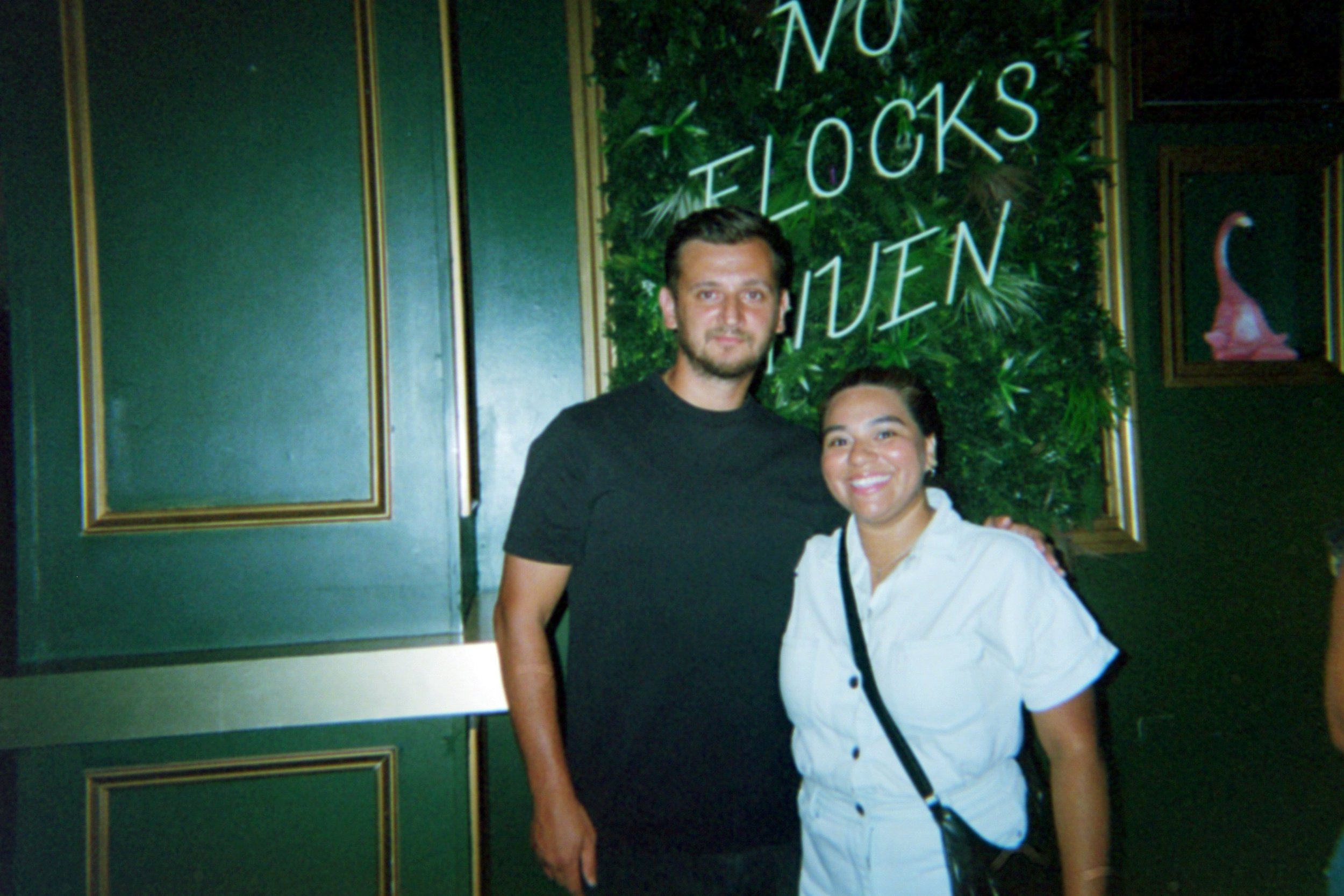A man and woman smiling and posing together indoors in front of a decorative wall with neon lights that spell 'No F**cks Given' and green foliage.