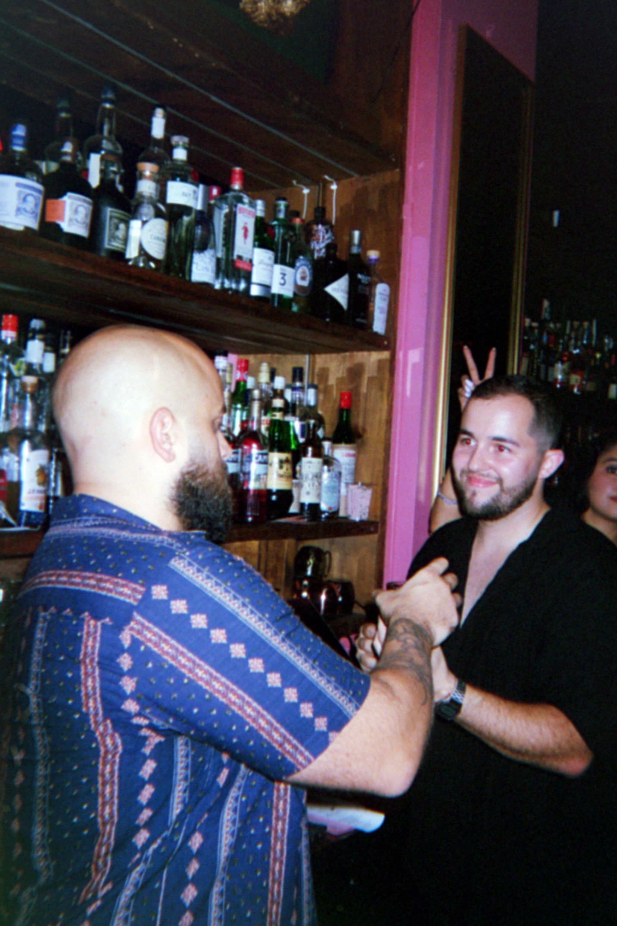 Two men in a bar. One has a bald head and beard, wearing a patterned shirt, and the other has dark hair and a beard, wearing a black shirt. The man in black is smiling, and the man with a beard is holding his hand. The man in the background is making
