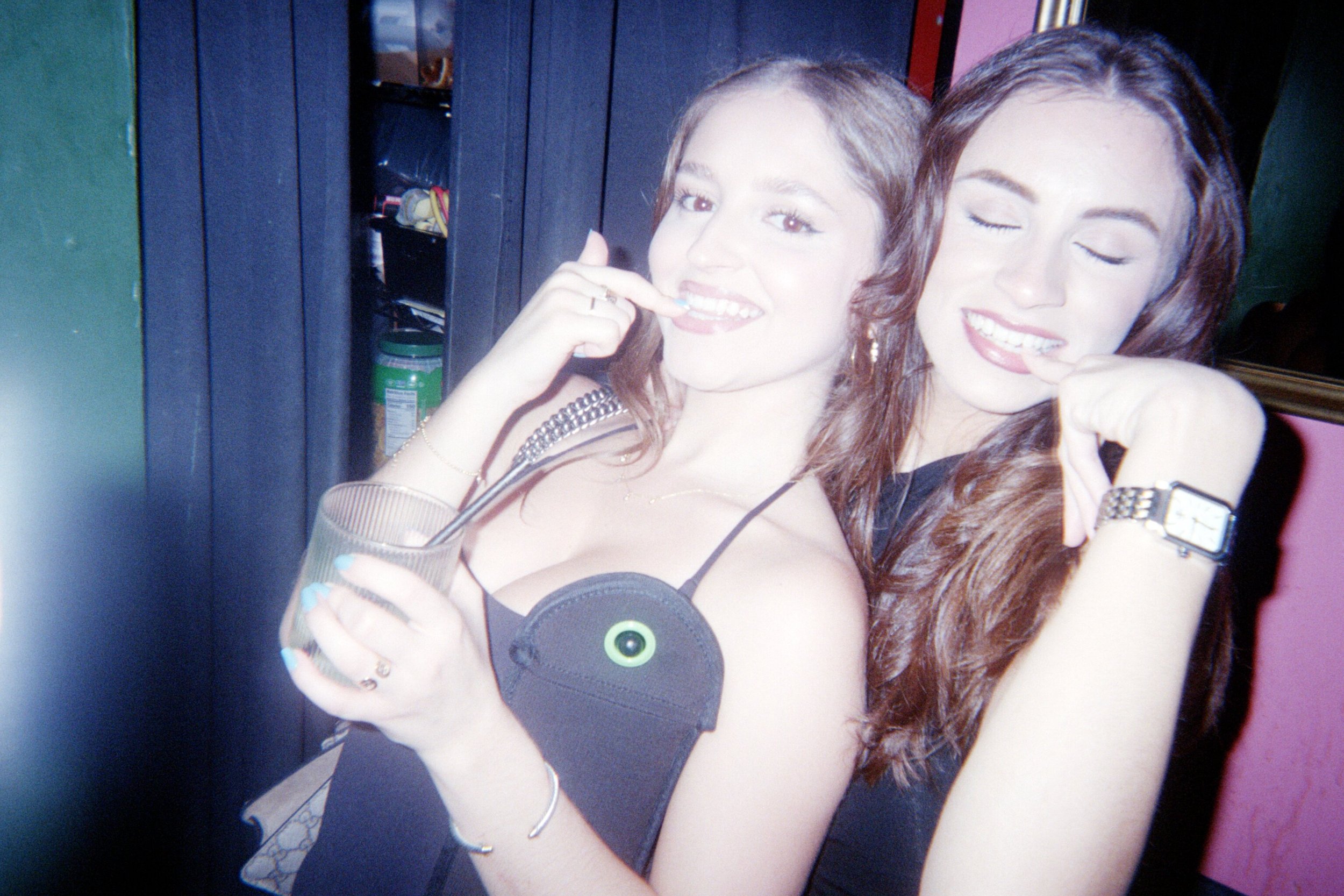 Two women posing close together at a party or club, one holding a drink with a straw and the other with a wristwatch, both smiling and making playful gestures.
