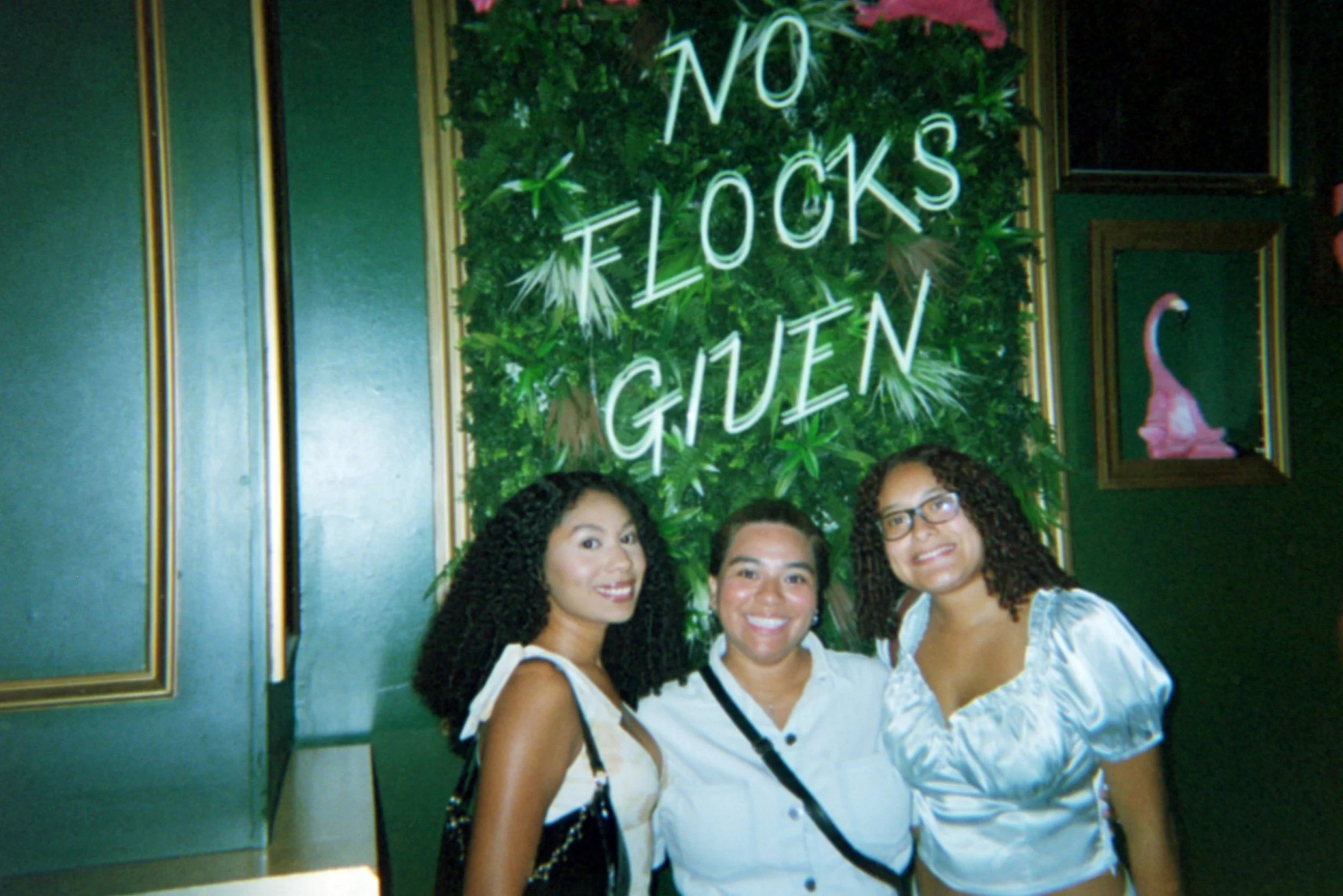 Three women smiling in front of a green leafy wall with a neon sign that reads 'No Flocks Giuen' and framed floral artwork on the wall.