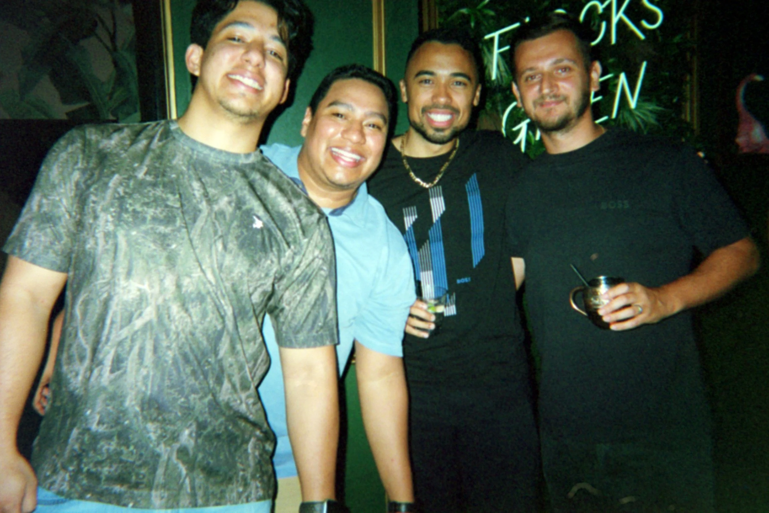 Four men smiling at a party, holding drinks, standing in front of a neon sign and a dark background.