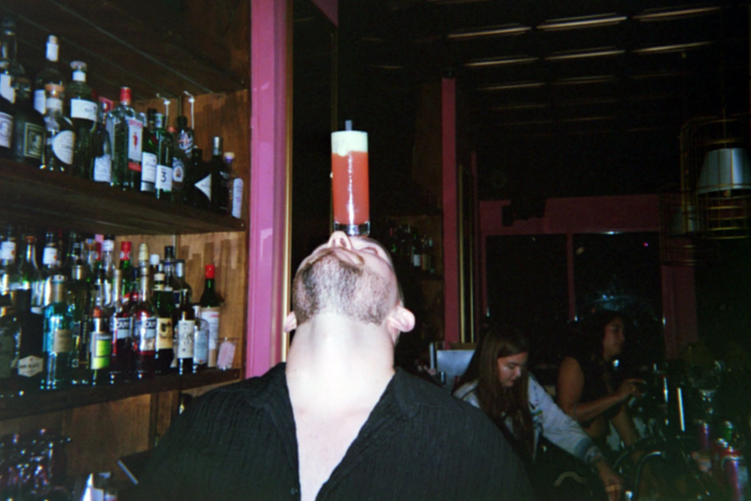 A man with a beard balancing a tall, layered red and yellow cocktail glass on his chin in a bar. Two women sit at the bar in the background, with bottles of alcohol on shelves behind them.