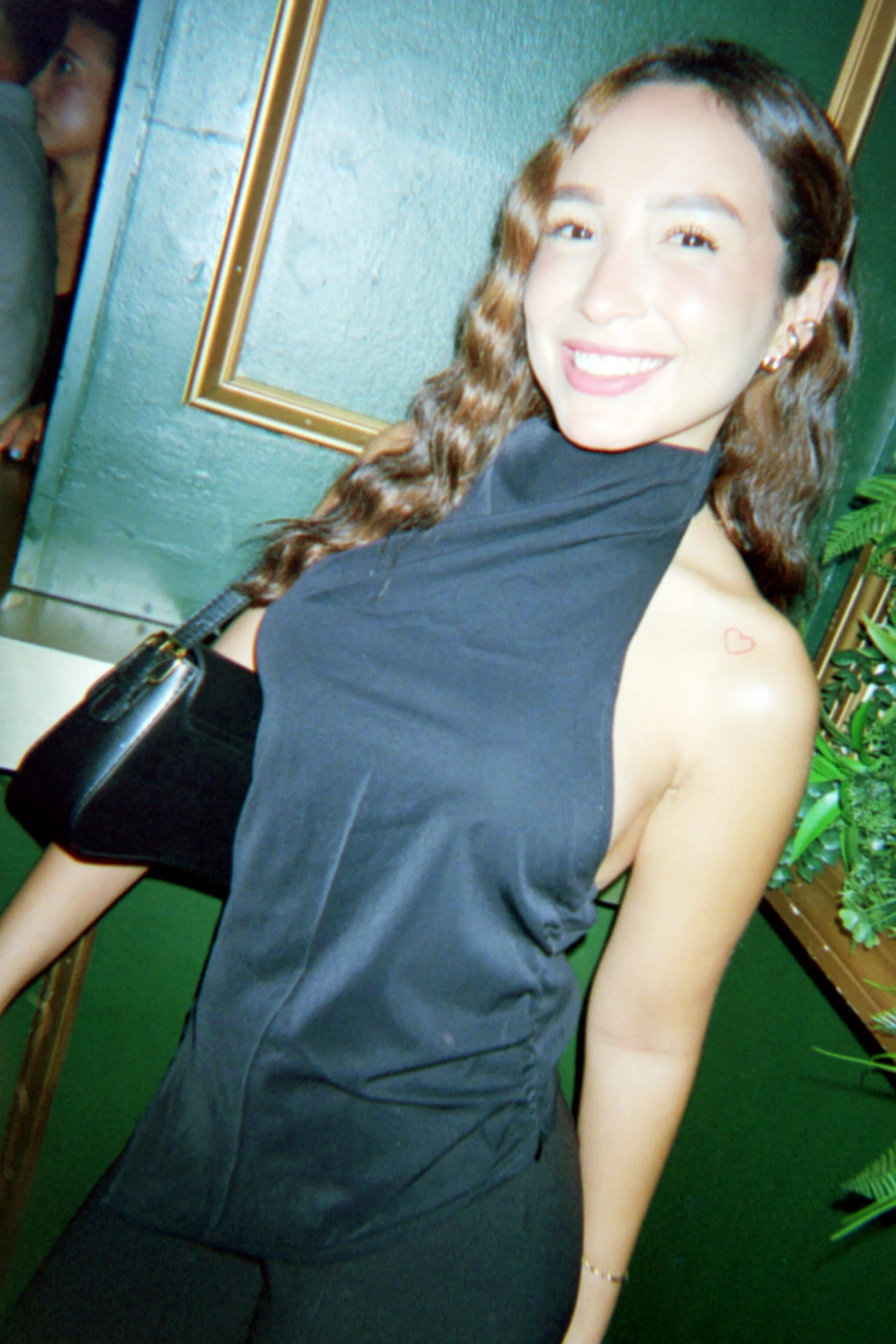 A woman with wavy brown hair smiling and looking at the camera. She is wearing a sleeveless black dress and has a small red heart tattoo on her left shoulder. She carries a black purse with a chain strap and is standing in front of a green wall with 