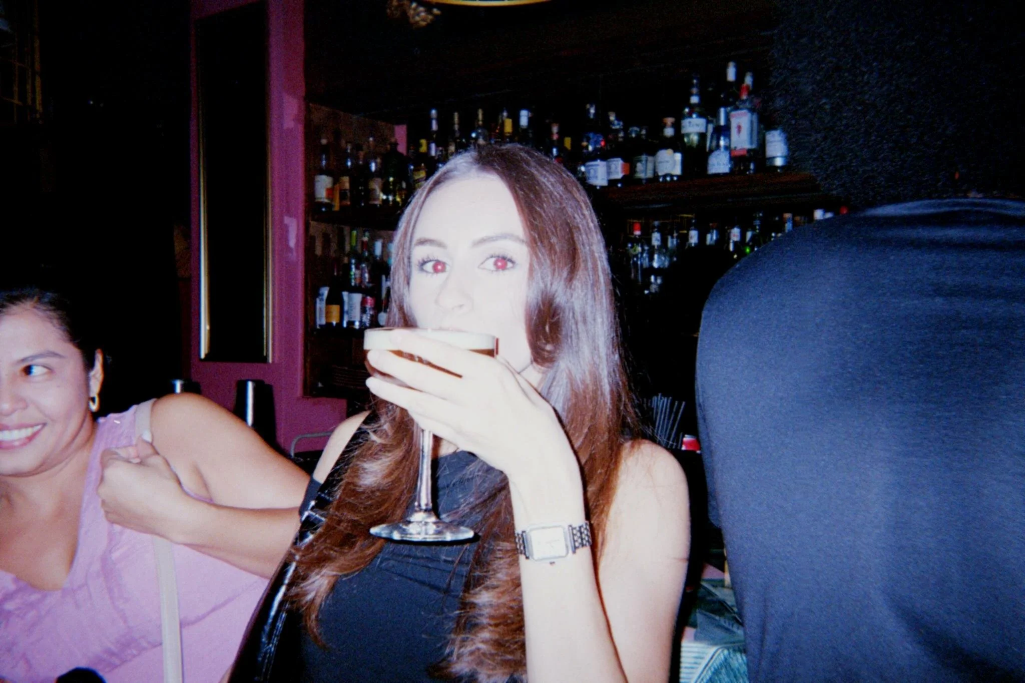 A woman with long brown hair drinking a dark beverage from a cocktail glass in a bar, with another woman smiling beside her, and shelves of liquor bottles in the background.
